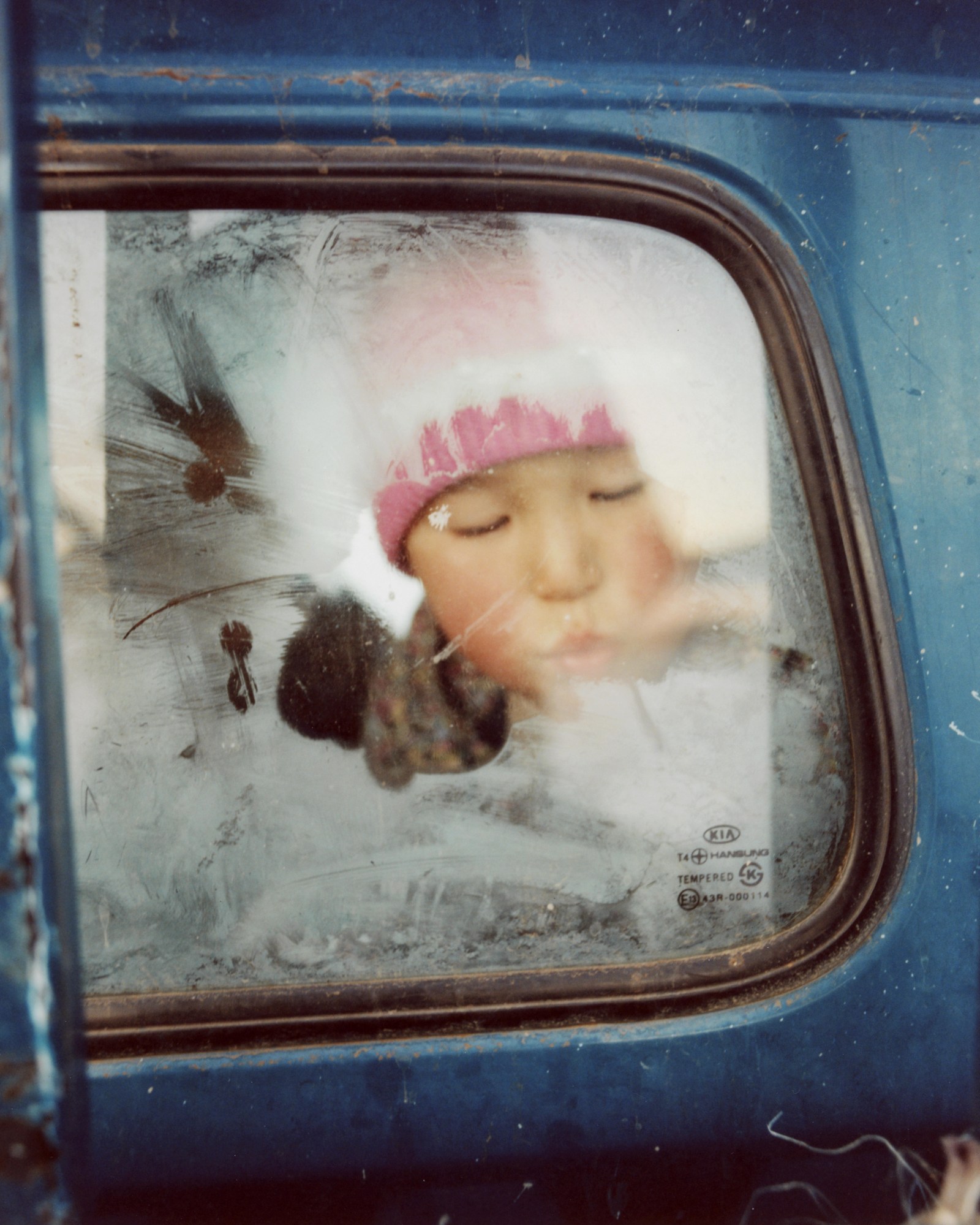 a young child in a pink hat breathing steam onto a cold car window