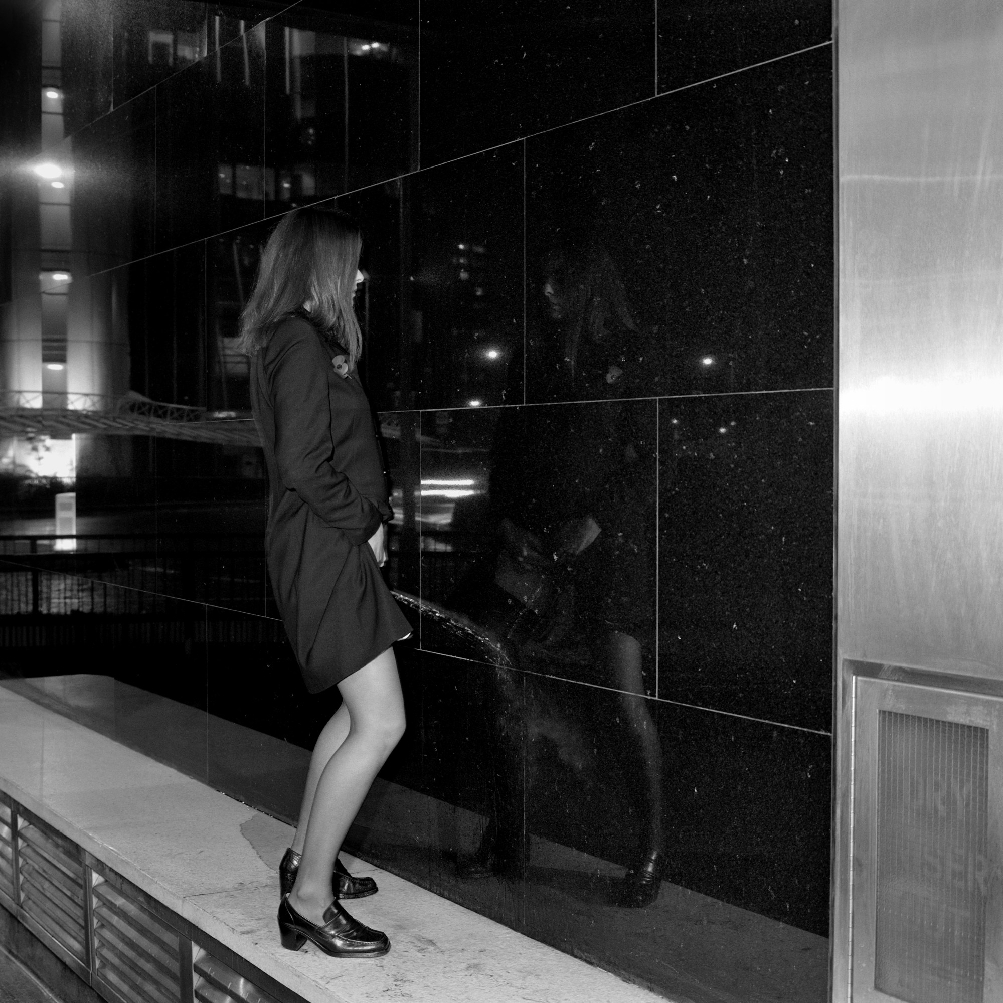 woman in black trench coat peeing on a reflective wall