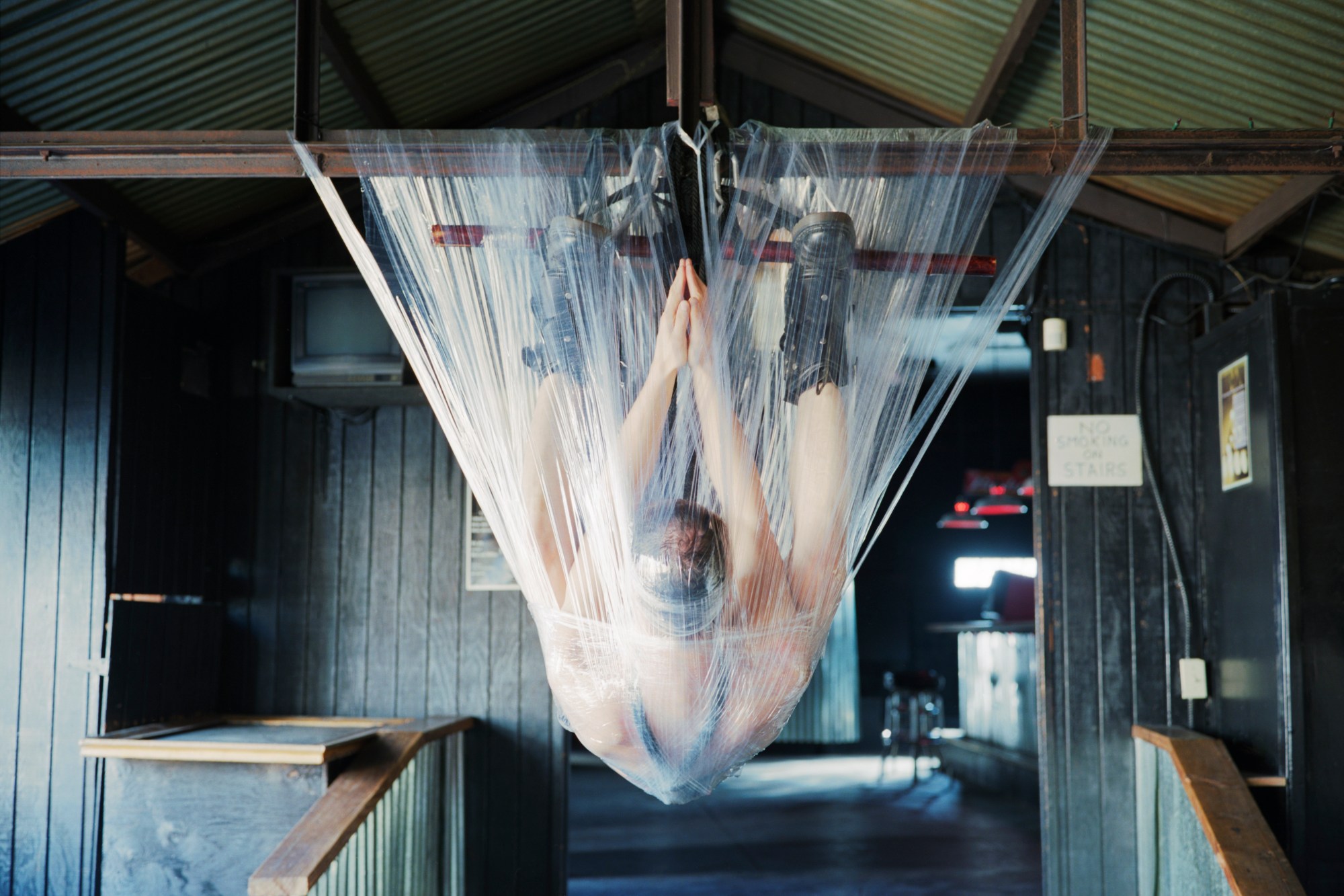 a person suspended from a ceiling beam by ropes and straps, covered in cellophane