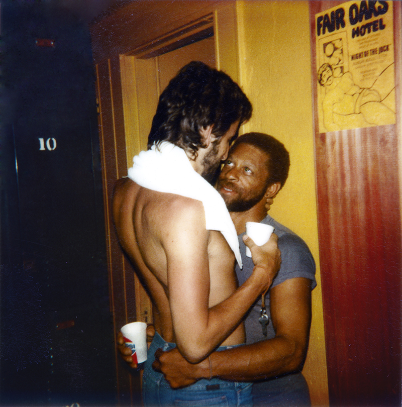 a black man sitting down wraps his arms around a white man standing in blue jeans shirtless above him.