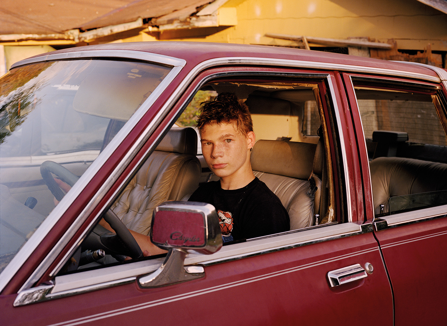 a teenage boy with freckles sits in an old burgundy coloured car, looking through the missing window to camera