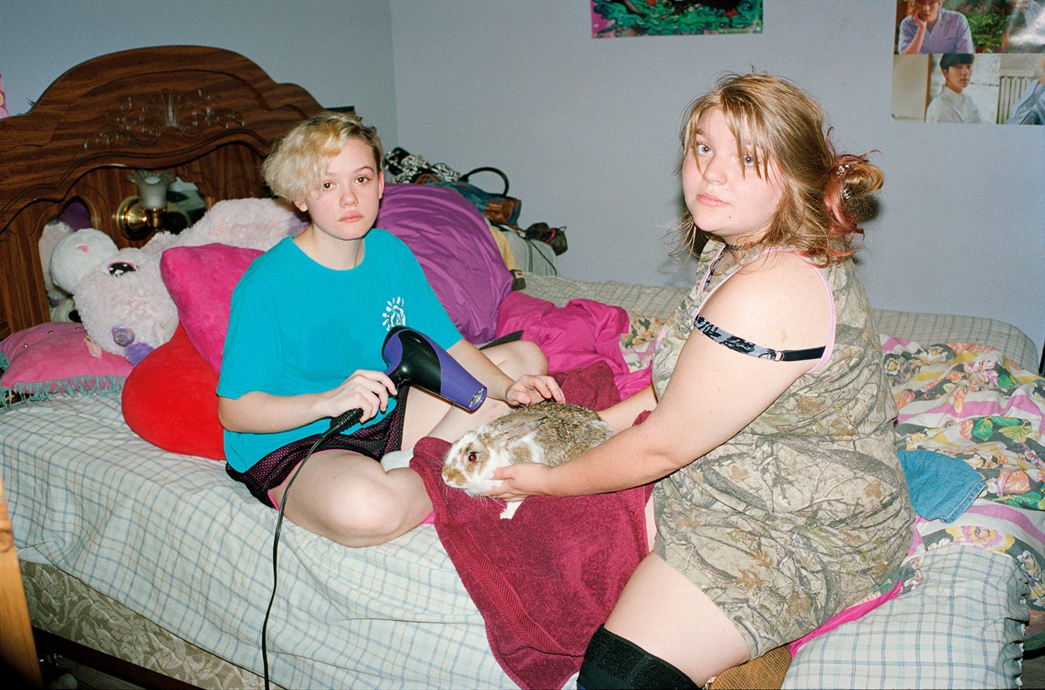 teen siblings sit on a messy bed using a hair dryer on their pet rabbit