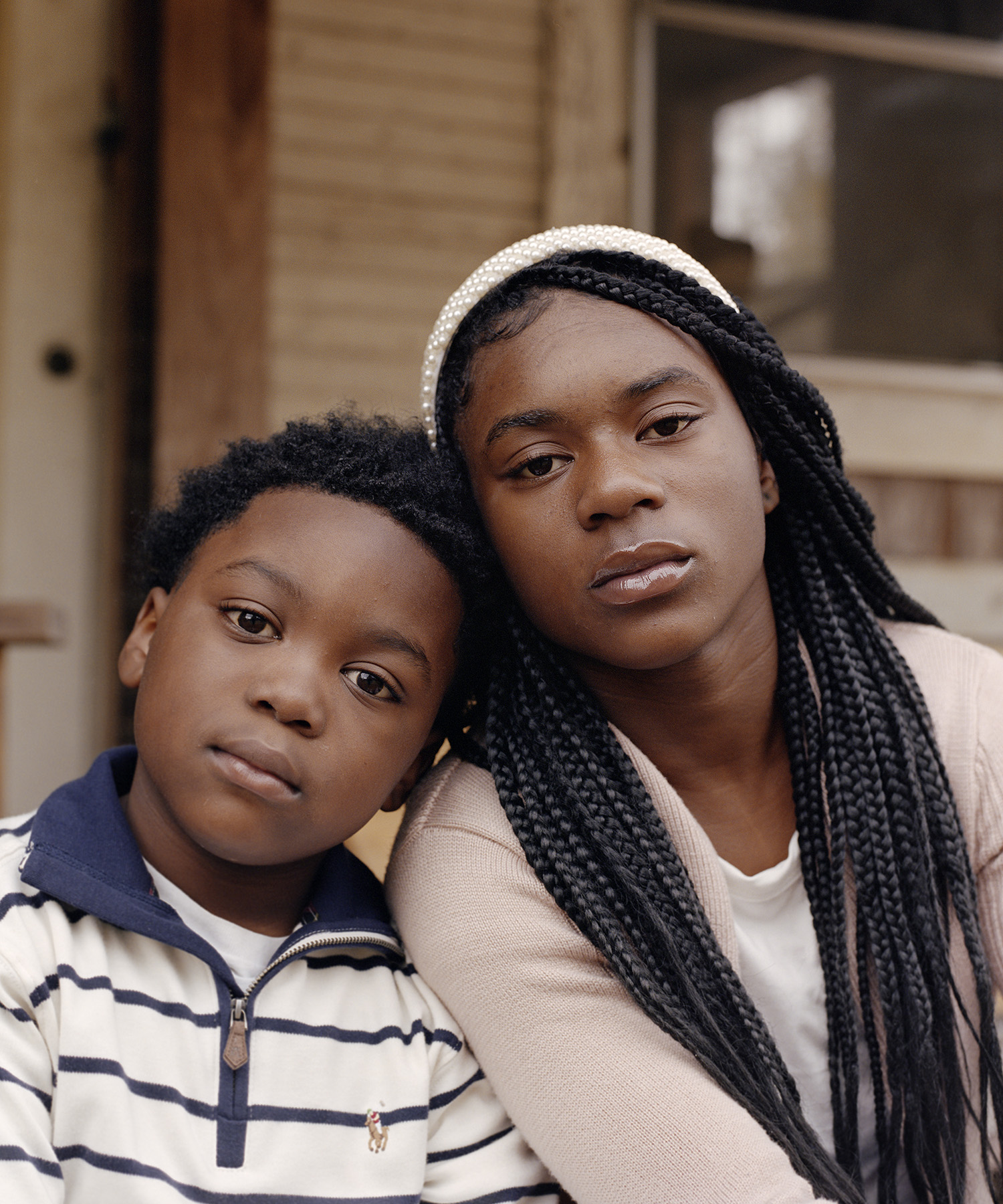 2 siblings sit side by side, their heads resting against each other, for a straight-faced family portrait