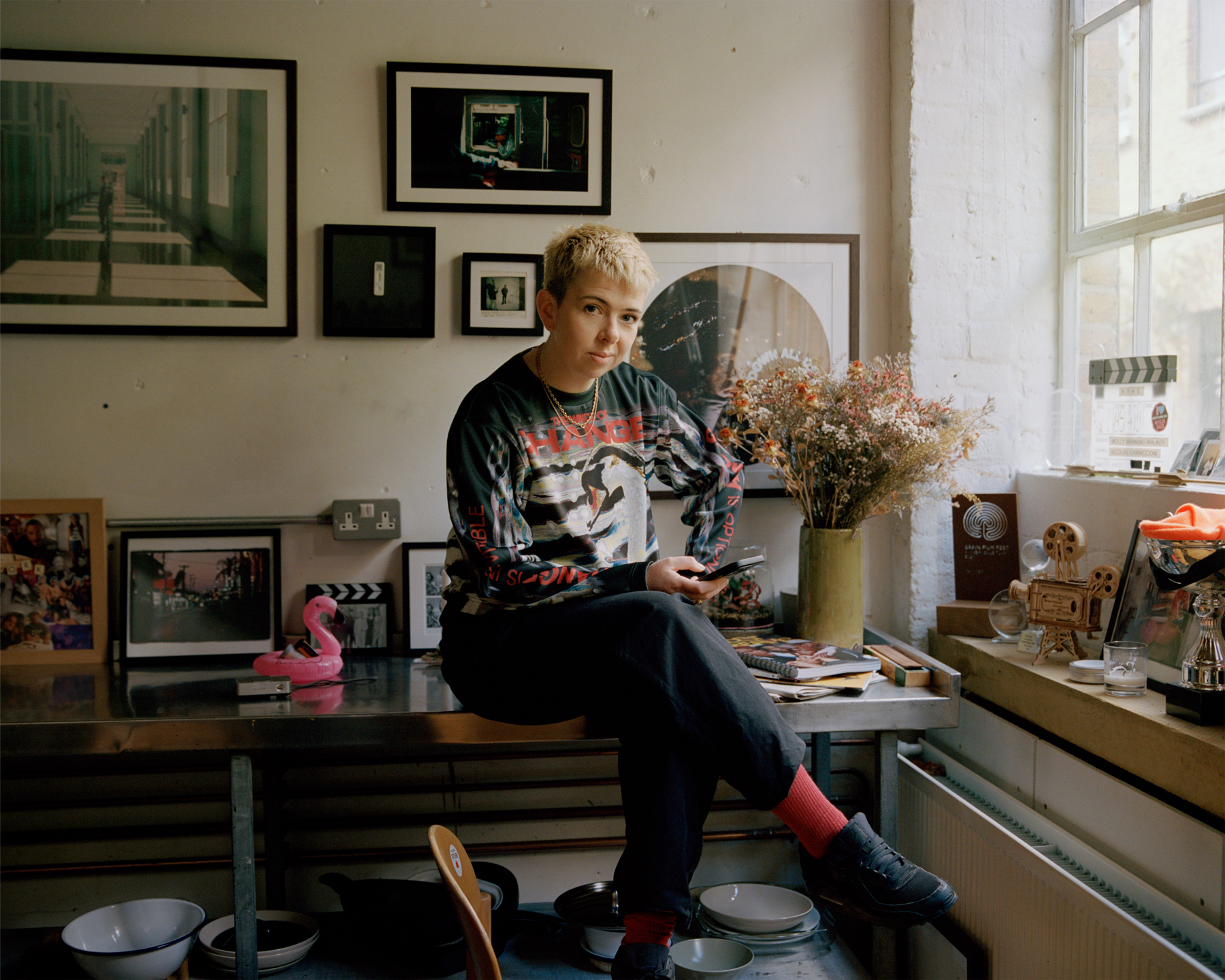 molly manning walker sitting on a stainless steel worktop in her kitchen surrounded by dried flowers, framed pictures and film awards