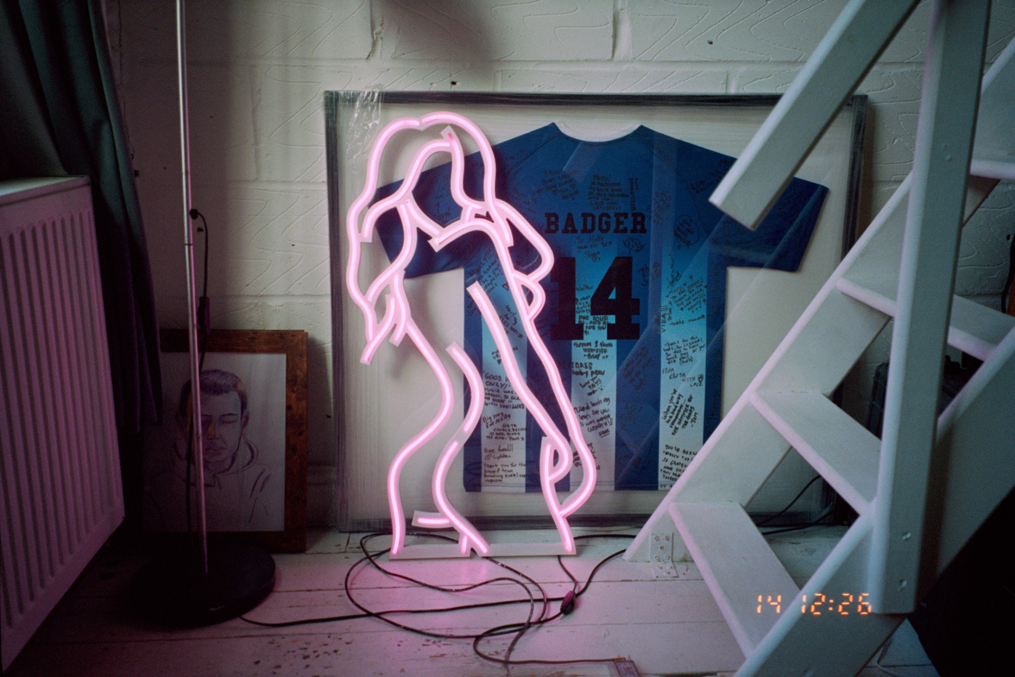 a corner of molly manning walker's apartment, with a framed and signed football T-shirt and a pink neon naked lady light