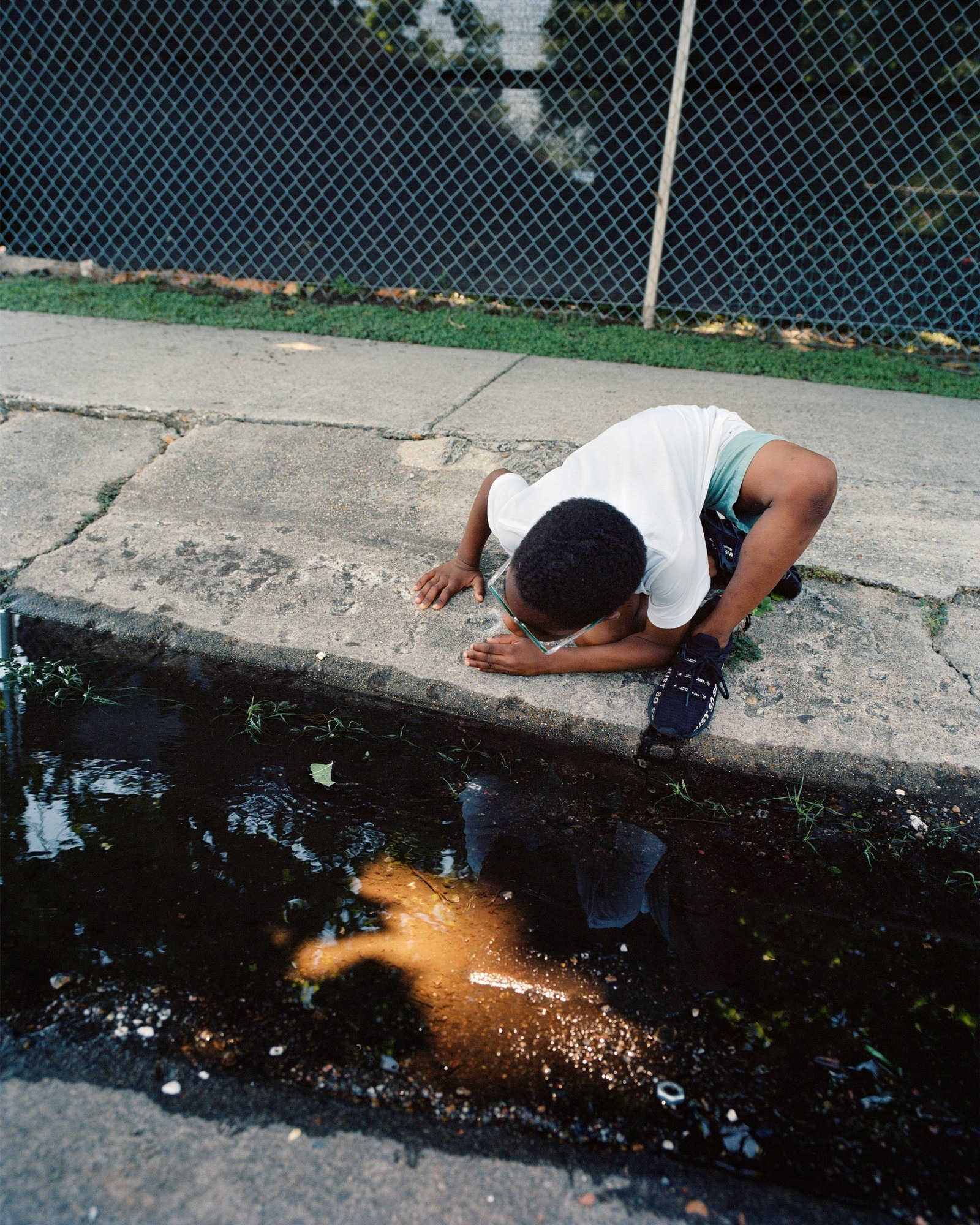 a kid knelt over a pavement looking a light reflection in a puddle