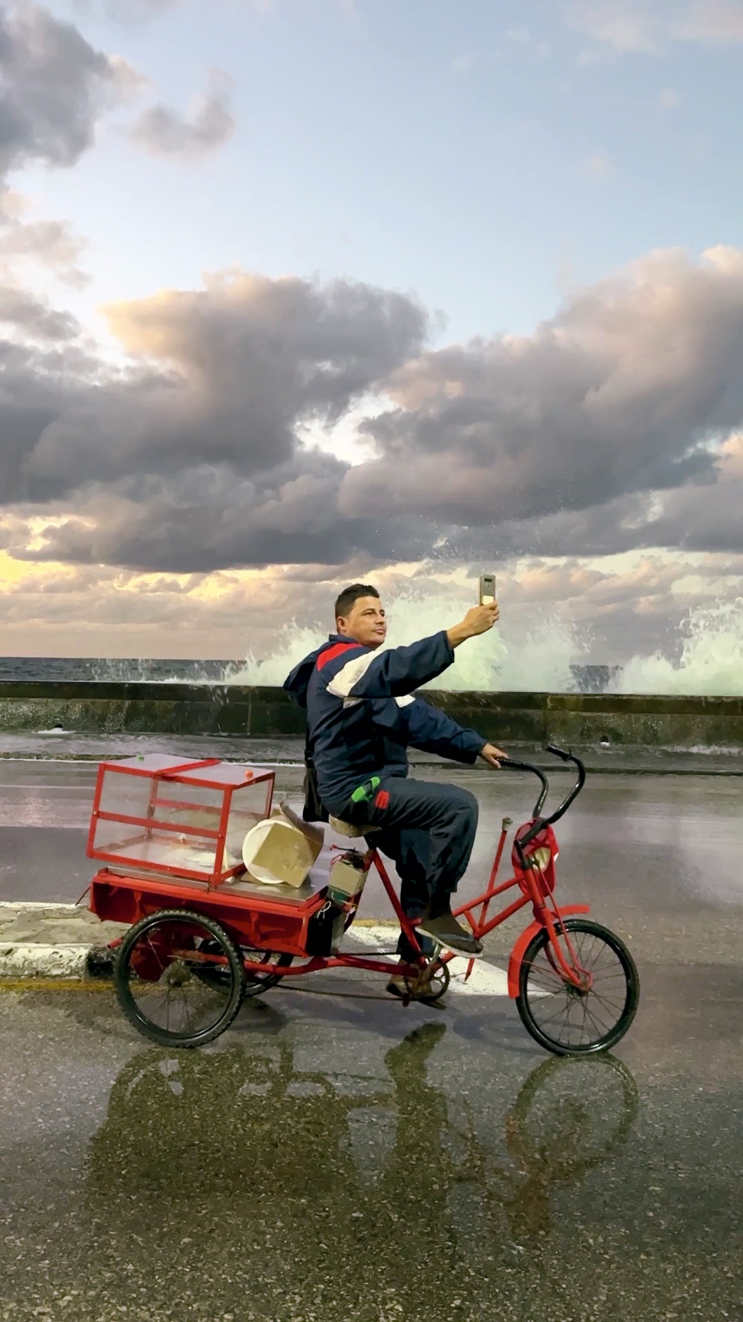 a man on a bicycle holding his camera phone up, the sea crashes in the background