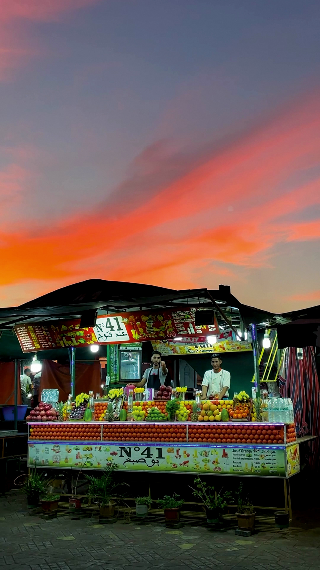 a neon lit stall with fruit and veg against a pink and blue sunset