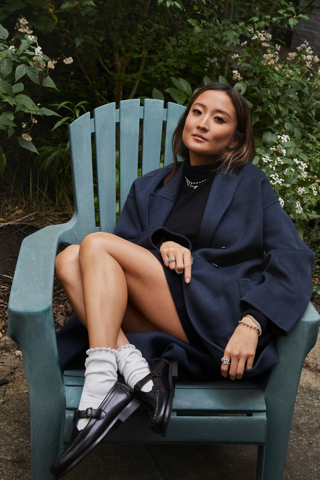 Ashley Park sitting in a green chair outside in a garden wearing black coat and shoes with white socks