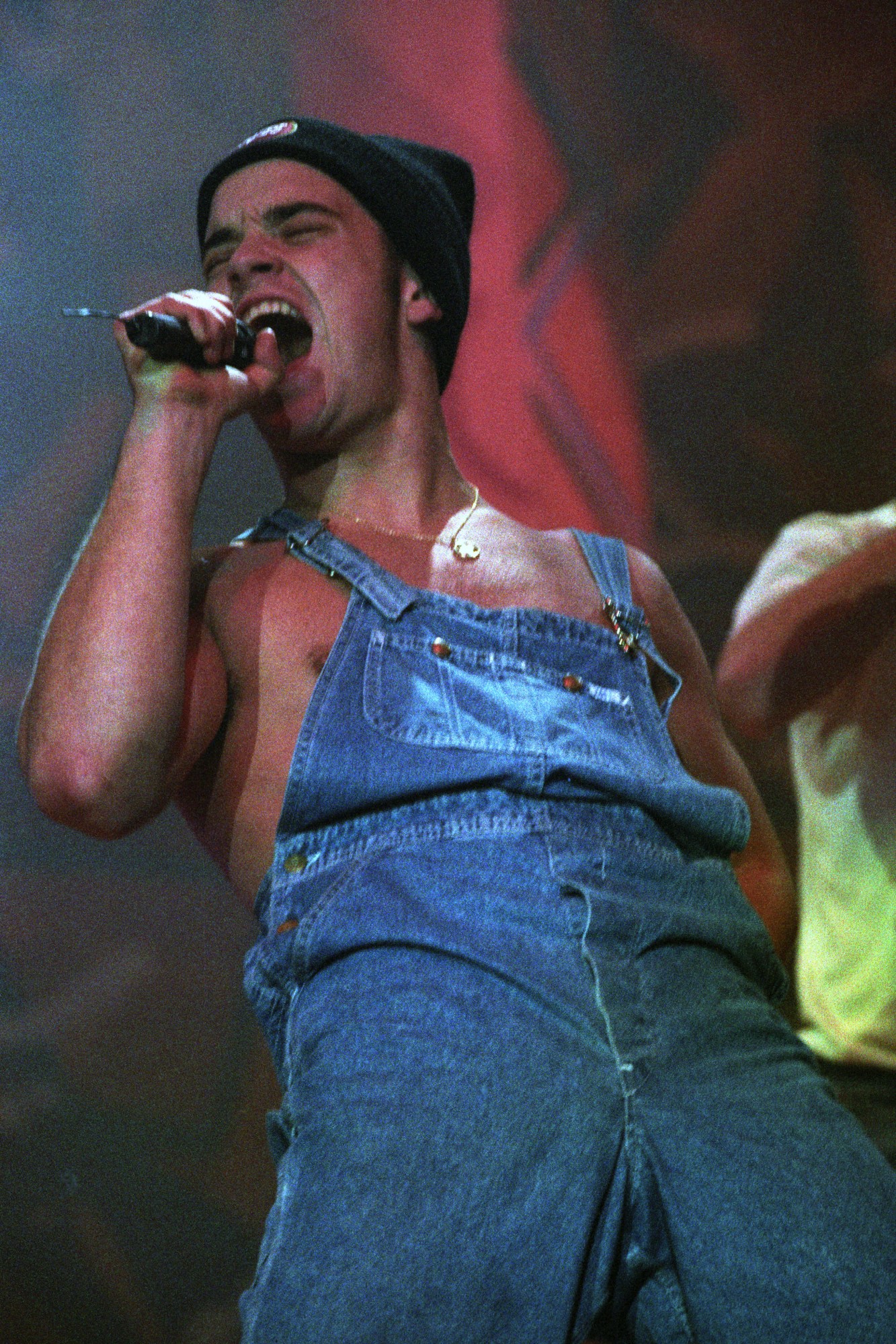 robbie williams performing in 1992 wearing a pair of denim dungarees with a beanie