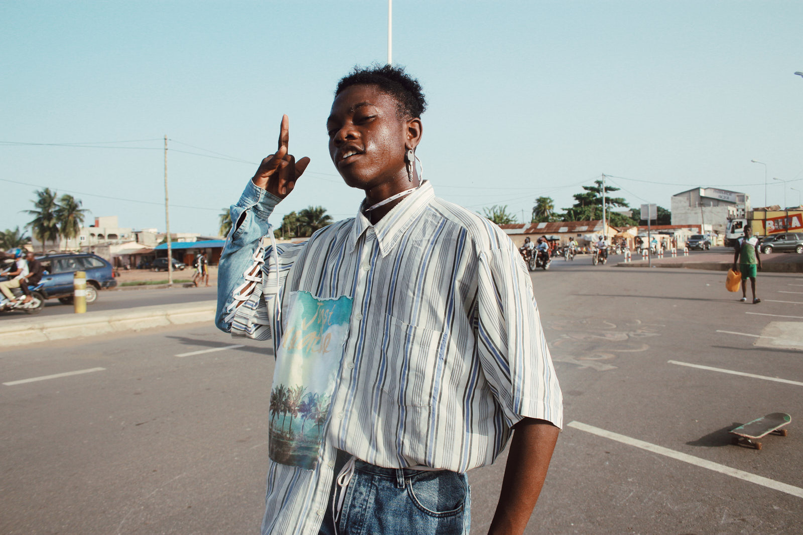 Patrick Arinzechukwu portrait of a person in a car park wearing a blue stripe shirt. they are black and have short hair and are sticking a finger up