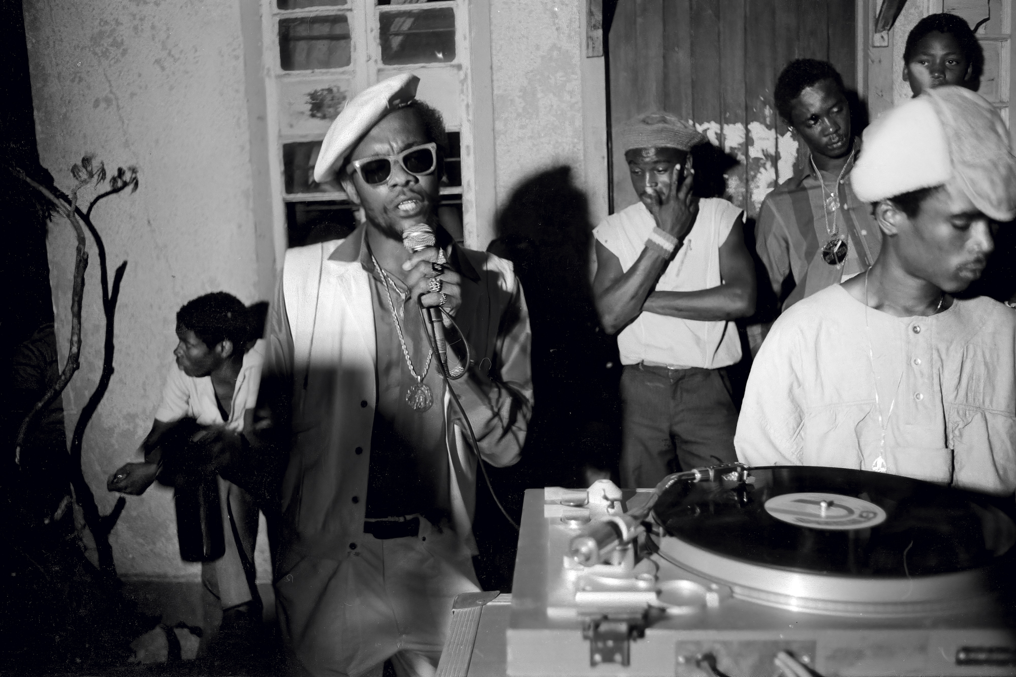 a man in a vest, beret and sunglasses singing into a microphone at a party in jamaica