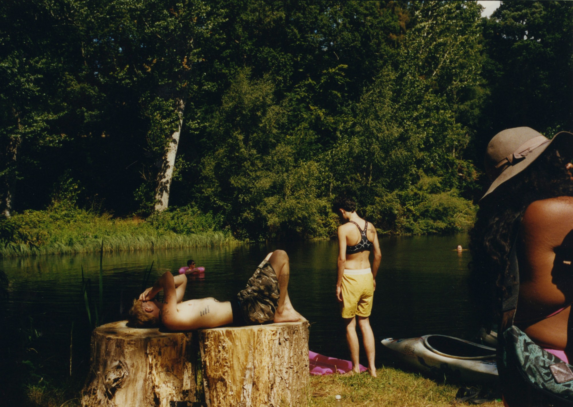 a topless person sunbathes on a large tree stump next to a picturesque lake; another two people are standing around nearby in swimwear