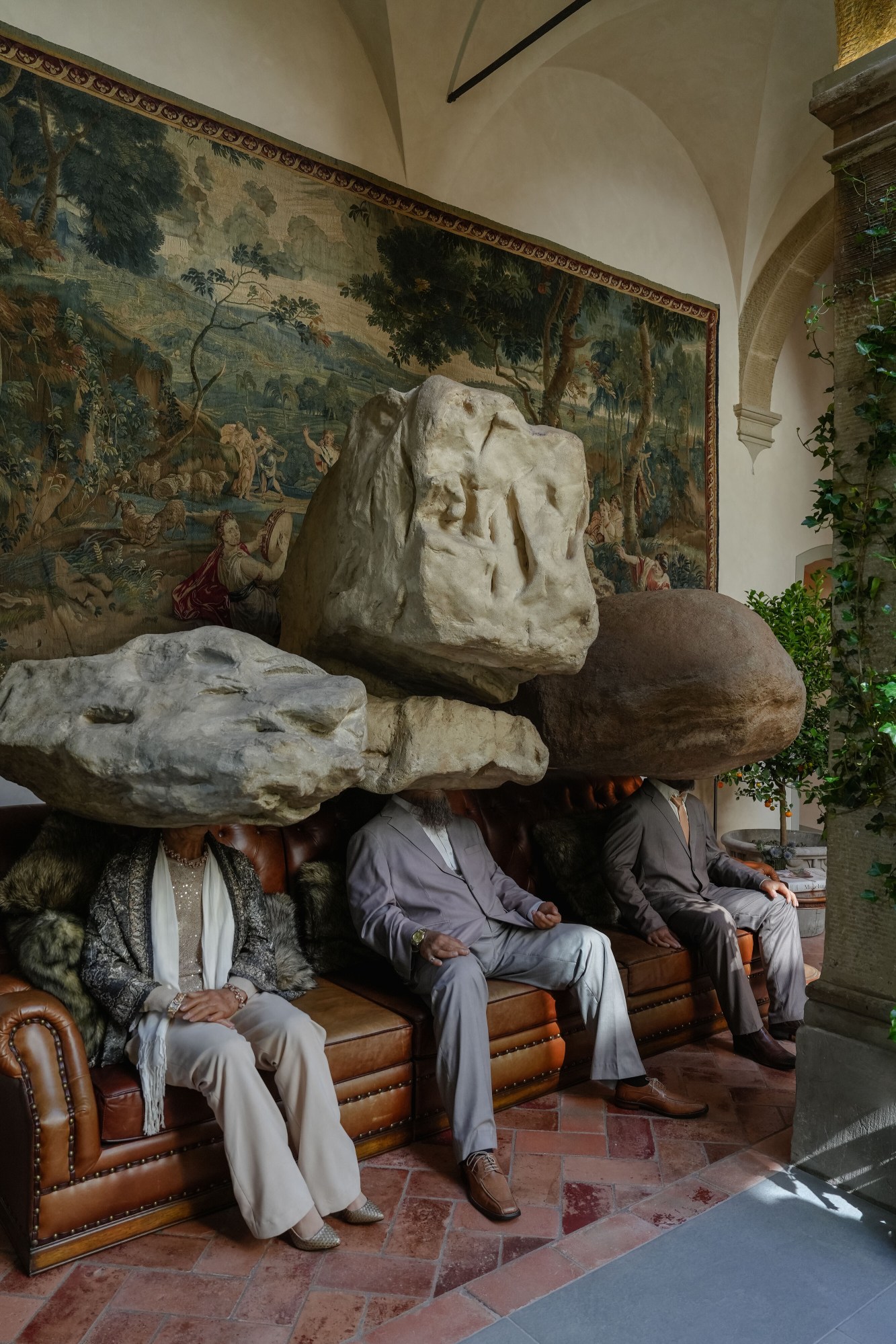 three lifelike statues with rocks for heads sit on a leather sofa in front of an elaborate tapestry