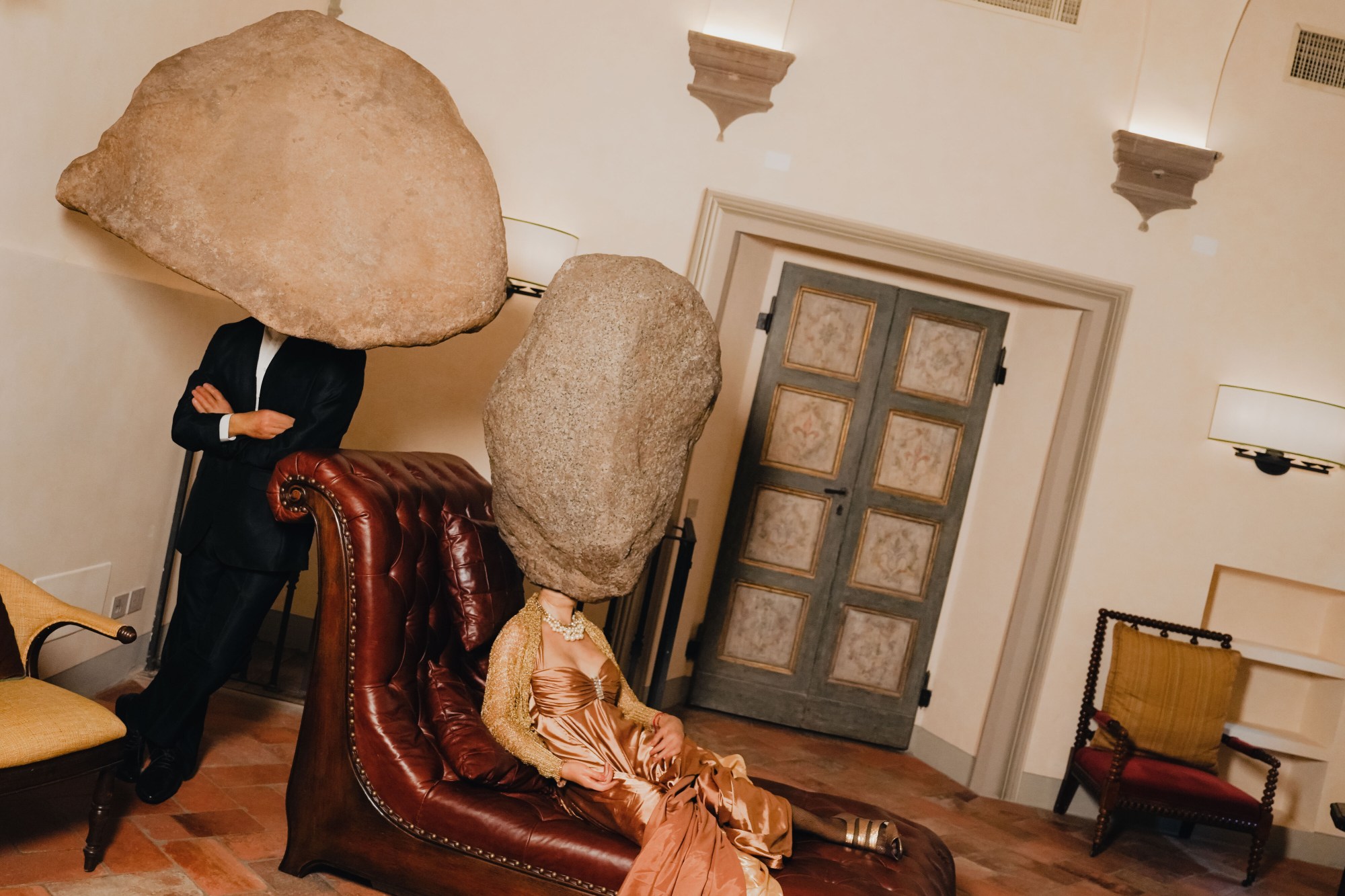 a statue of a woman in evening wear reclines on a leather chaise; a statue of a man in a suit leans against one end of it. both statues have big boulders for heads