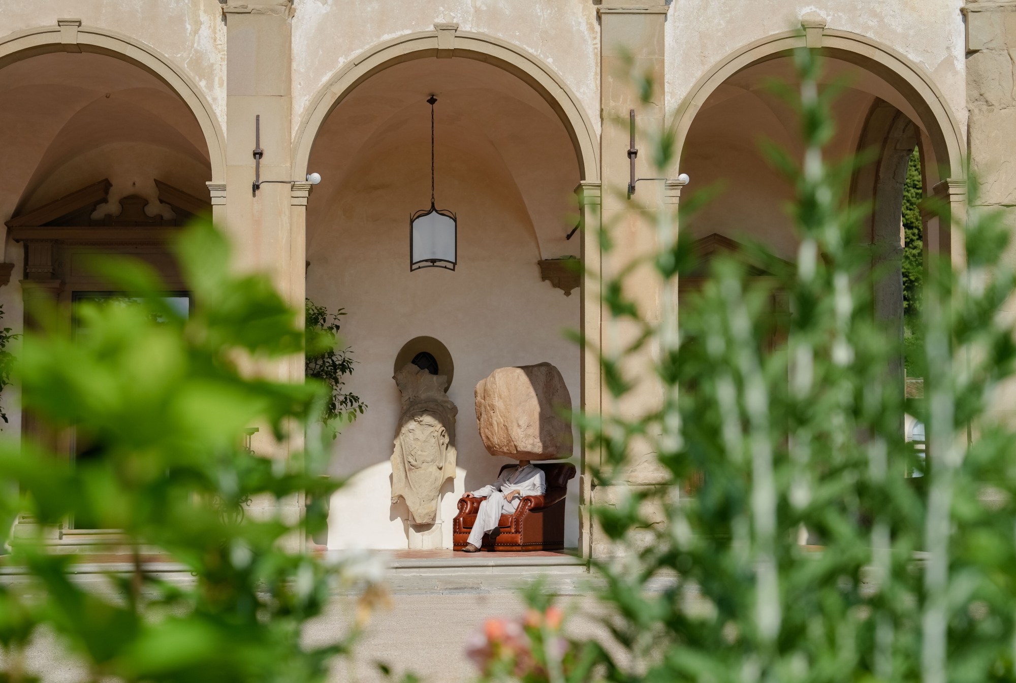 a statue of a man with a giant boulder for a head sits in a leather chair on a sunny terrace