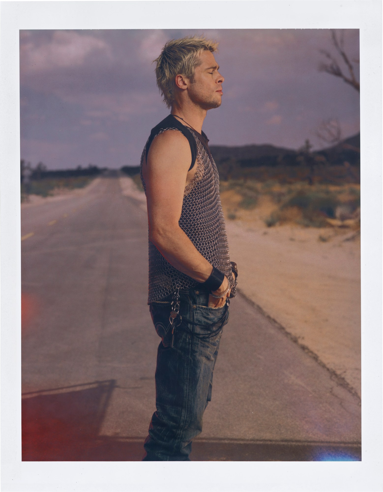 Brad Pitt in a chainmail vest and blue jeans, standing in profile on an empty dirt road