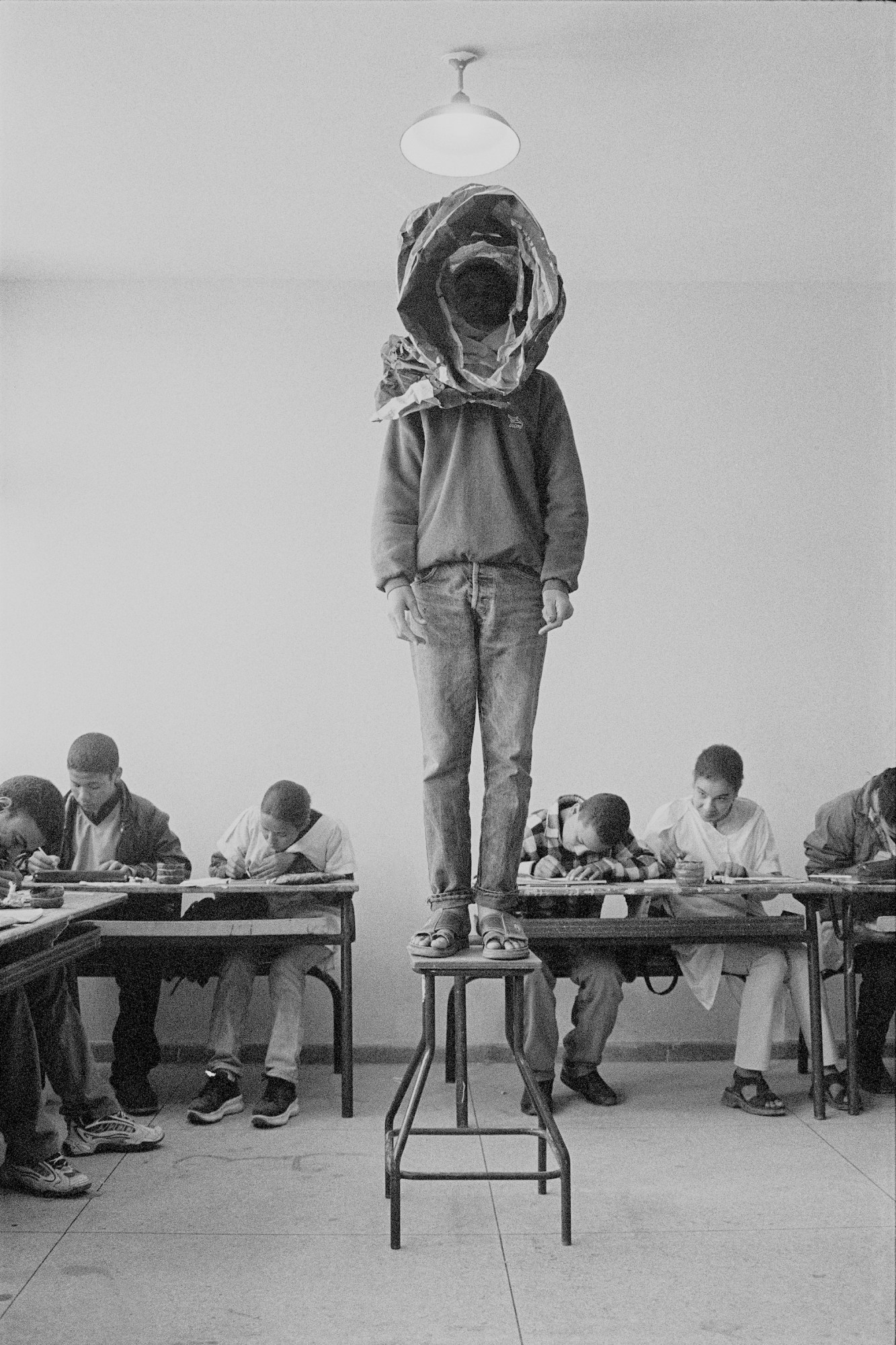 black-and-white photo of a boy standing on a desk in the middle of a classroom