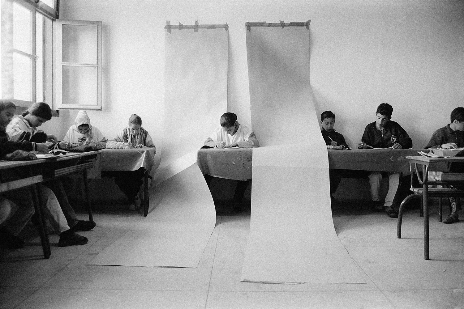 black-and-white photo of students in a classroom, long white curtains hanging over one desk