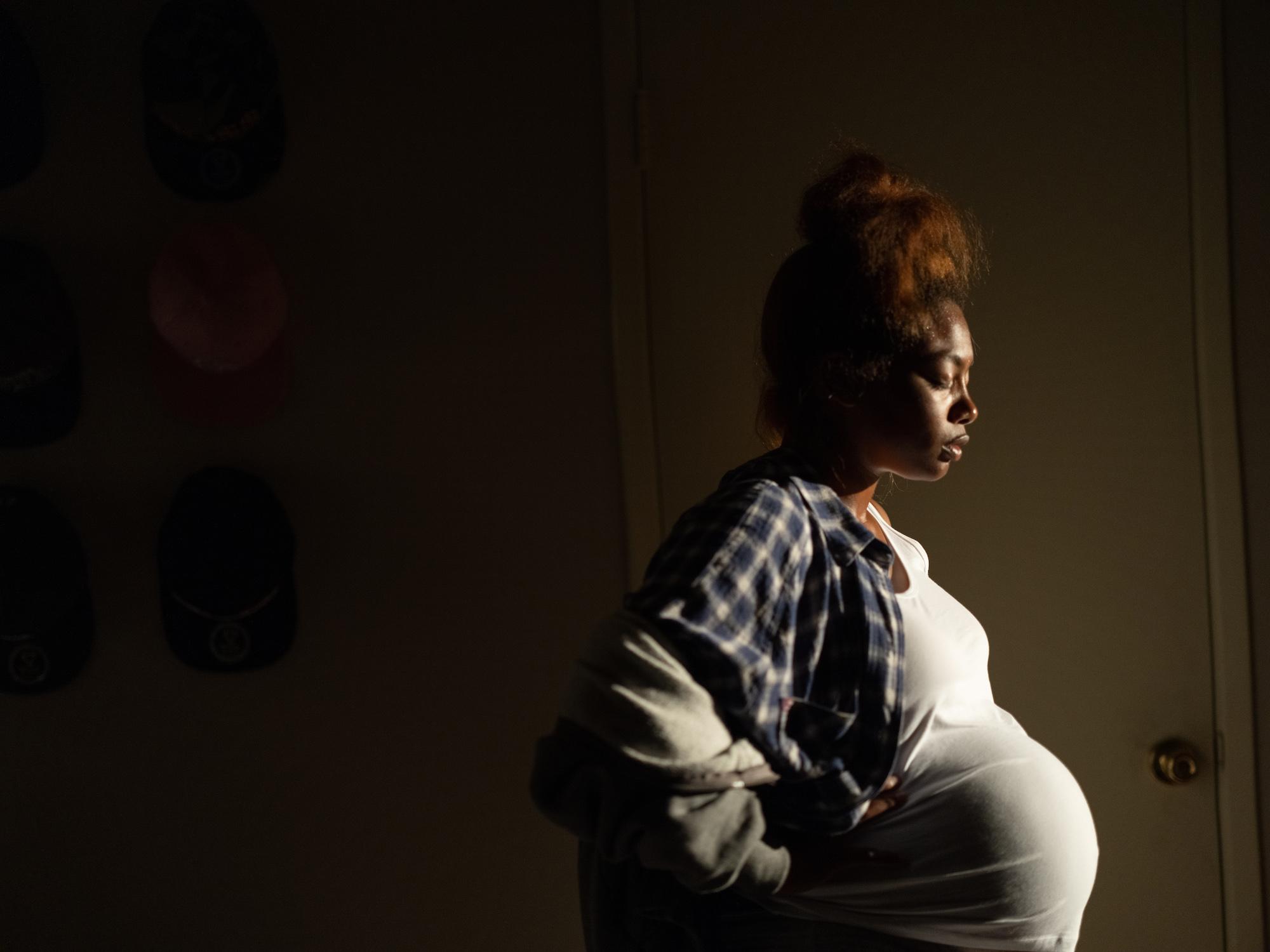 a pregnant young woman in a flannel shirt and white tank top standing in a dimly lit hallway