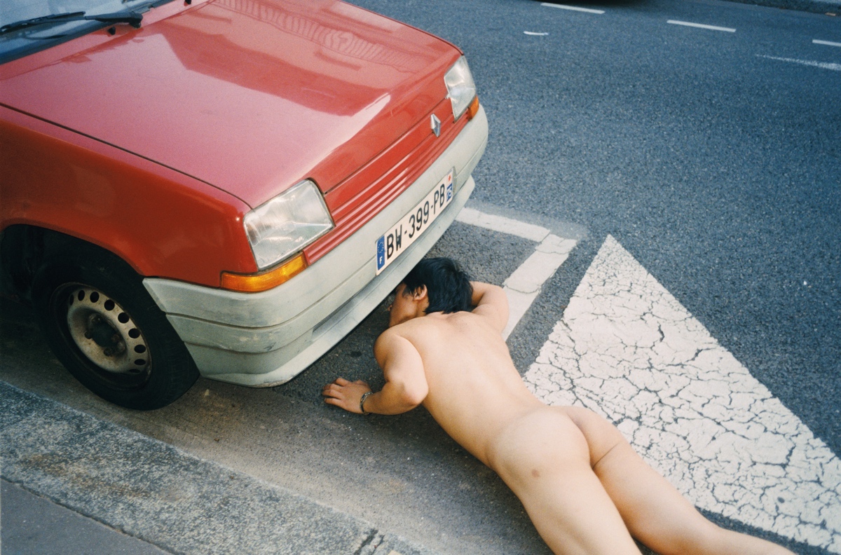 a naked man lies face down on the road looking underneath a red car