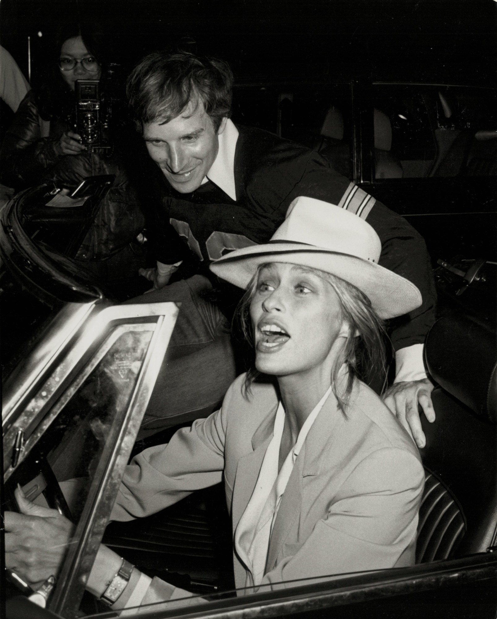 lauren hutton wearing a ferdora behind the wheel of her car, yelling at the papparazzi surrounding her