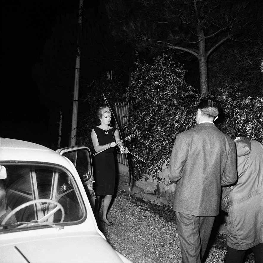 the actor anita ekberg stands on a neighbourhood sidewalk, pointing a prop bow and arrow at the paparazzi pursuing her