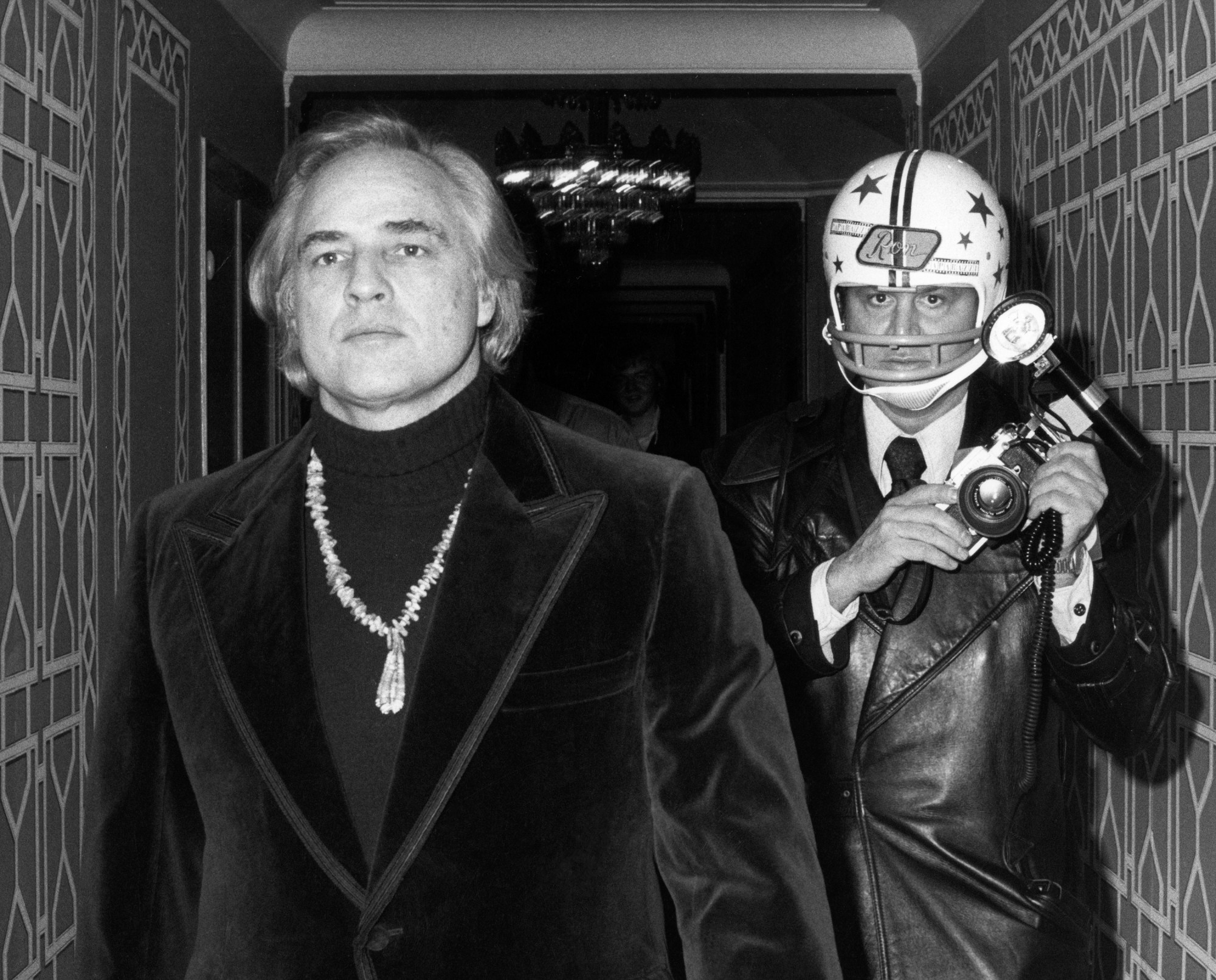 marlon brando walking down a corridor wearing a dark velvet suit, roll neck sweater and elaborate necklace; a photographer in a leather jacket and baseball hat walks alongside him