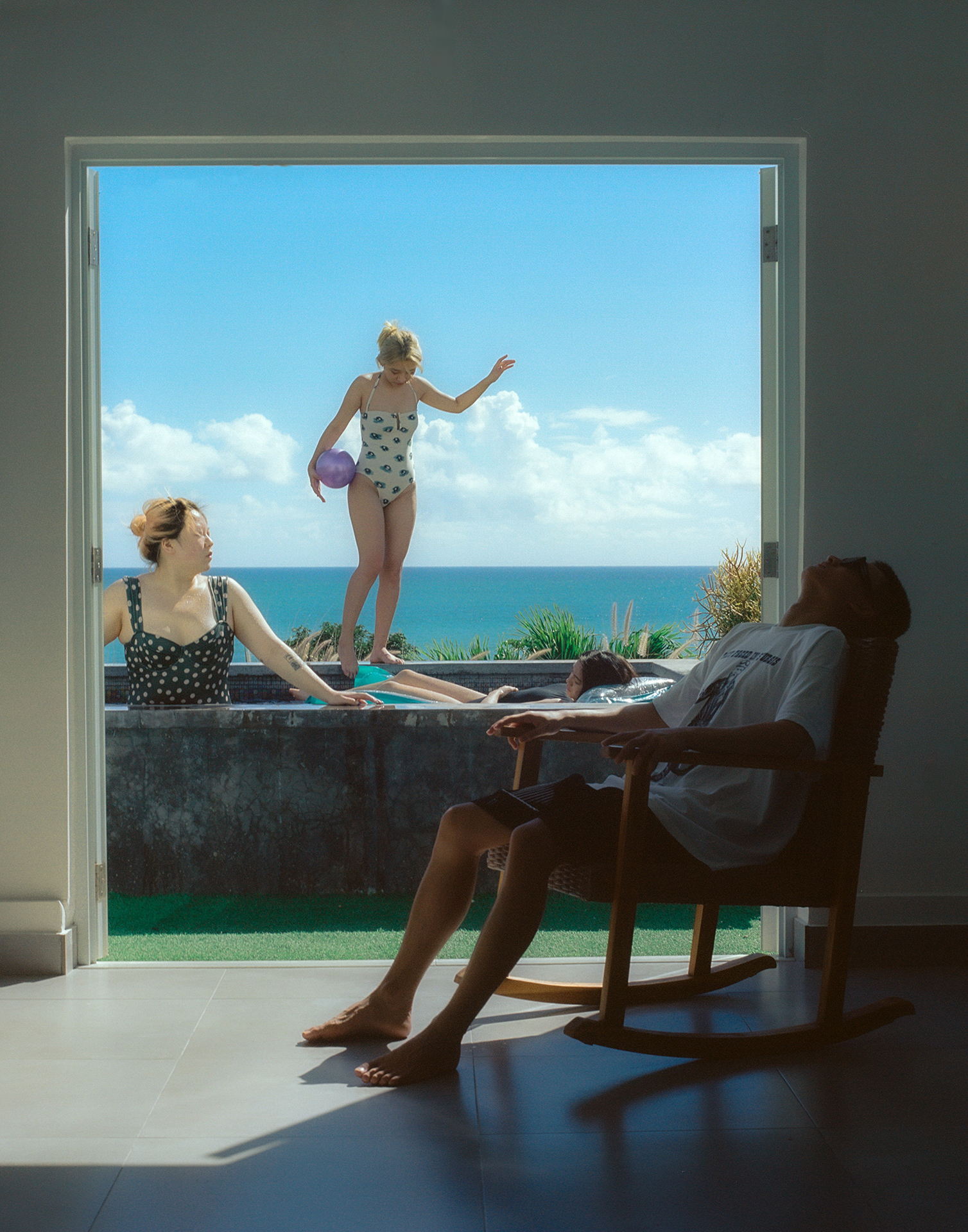 a group of friends look laze in a small pool with the sea behind them. in the foreground, a person sits on a chair in the shade, their head tipped back