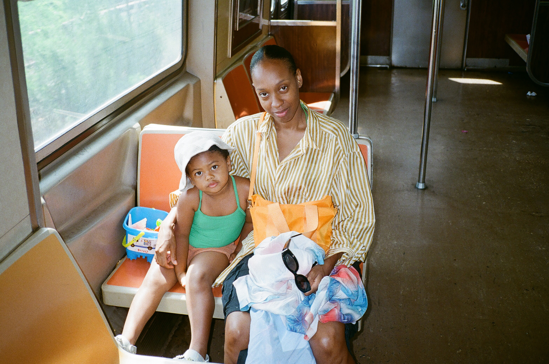 a woman in a striped shirt sits on public transport, her arm around a girl wearing a green t shirt and a white hat