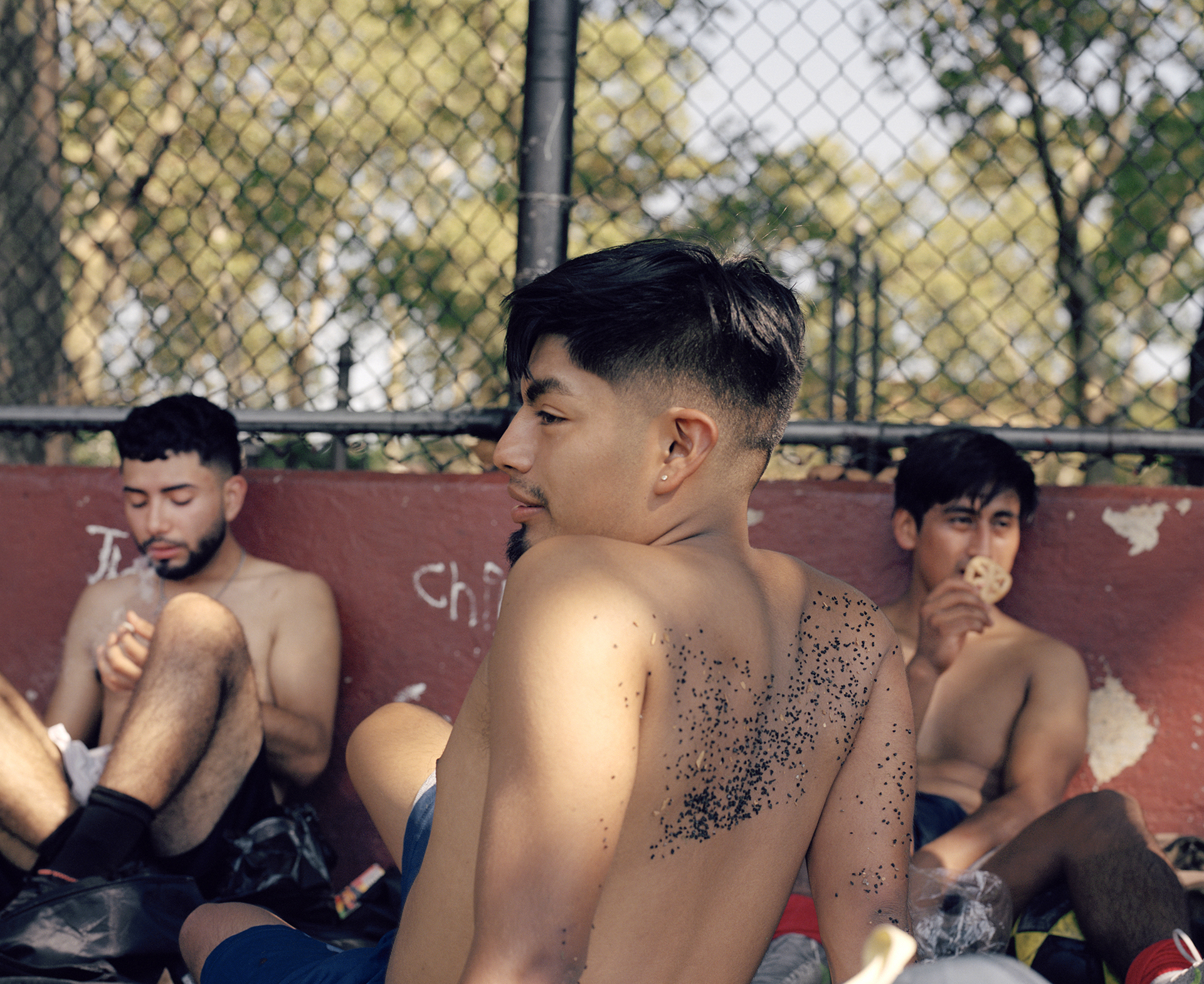 a group of three friends sit by a basketball court fence, one in the foreground sits with his back to us. to his left a boy smokes, on his right. a boy eats pretzels