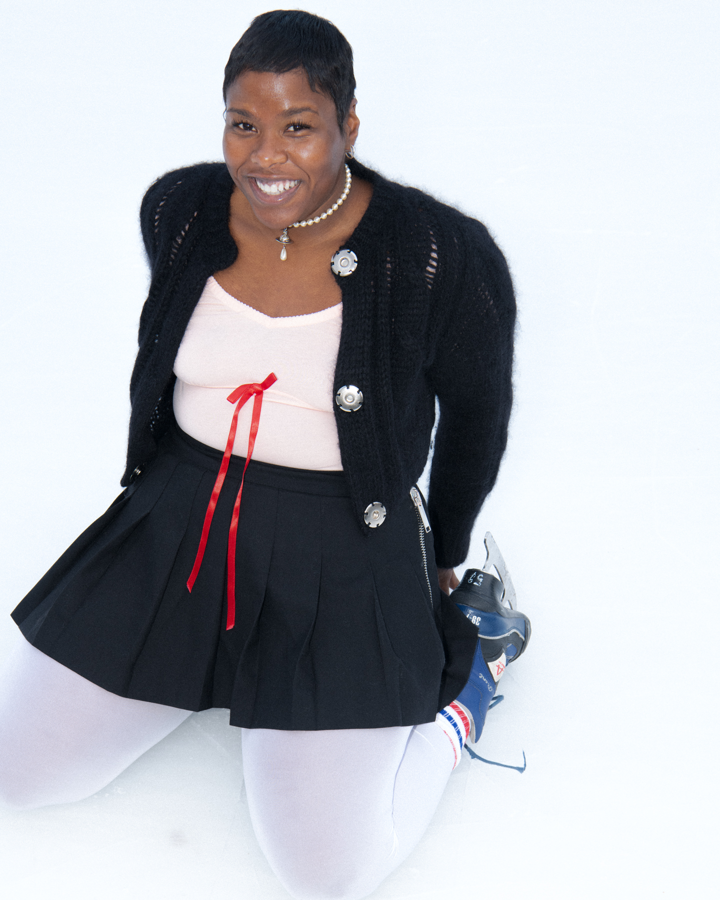 the artist precious okoyomon kneels on an ice skate rink wearing a skirt and cardigan; they smile at the camera