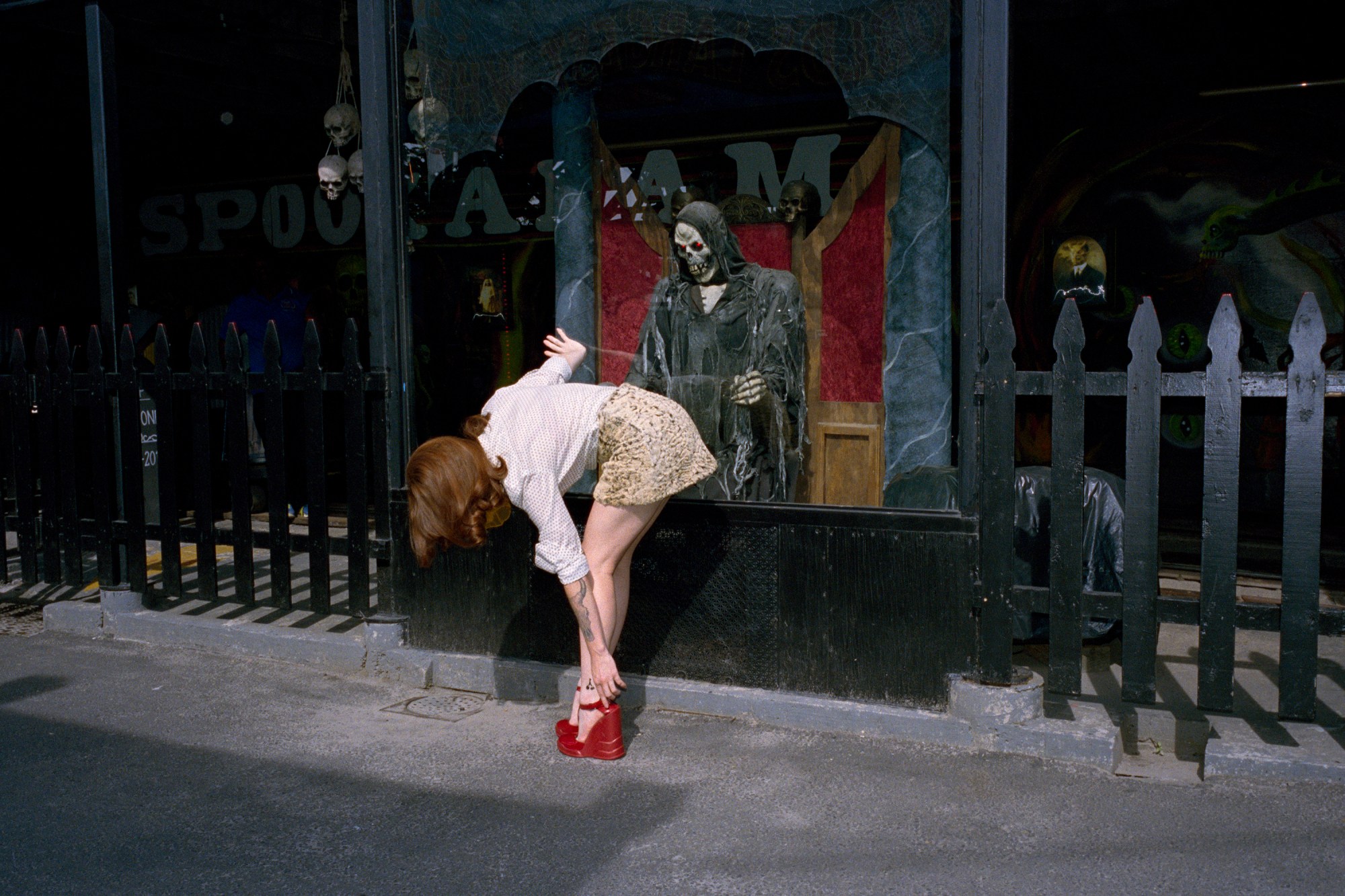 a daniel arnold photo of a redhaired women in red stilettos bending down to refix them. behind her in a glass box is a animatronic grim reaper, appearing to watch her.