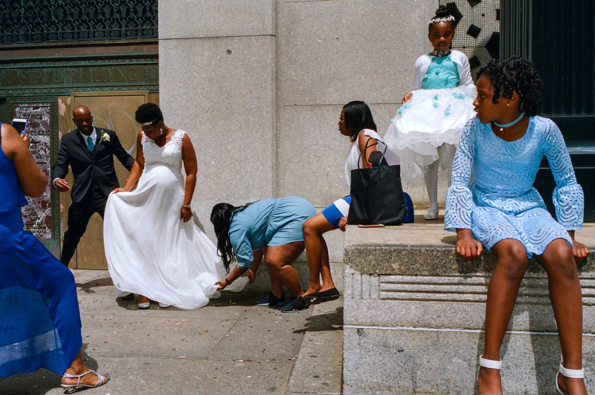 a family gather outside of new york city's clerk office, a new husband and wife has just got married and they're fixing the bride's dress. children dressed in shades of blue surround them.