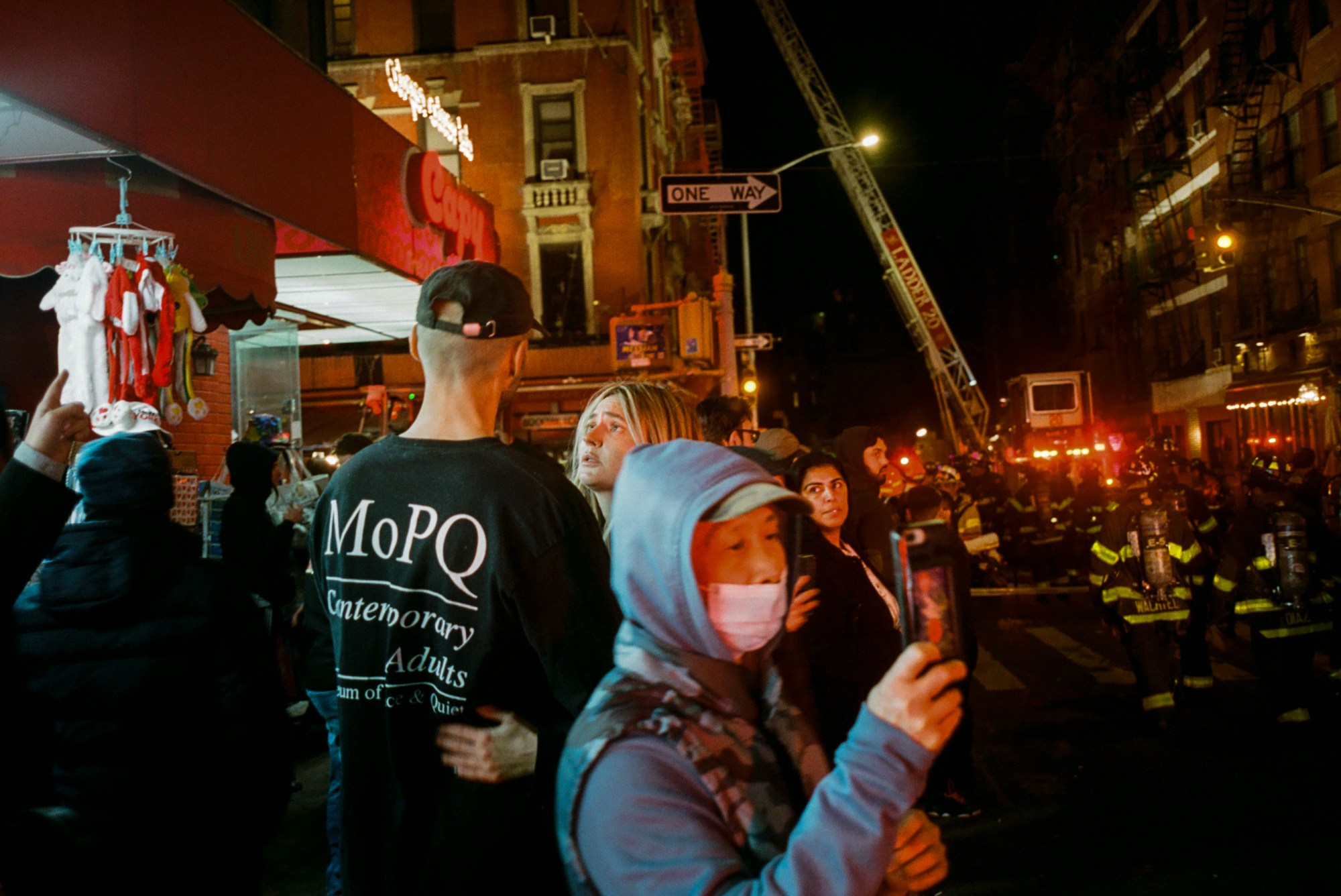 a group of people gather on a new york city street at night. a couple talk. a man wearing a mask takes a photo. in the background fire fighters gather.