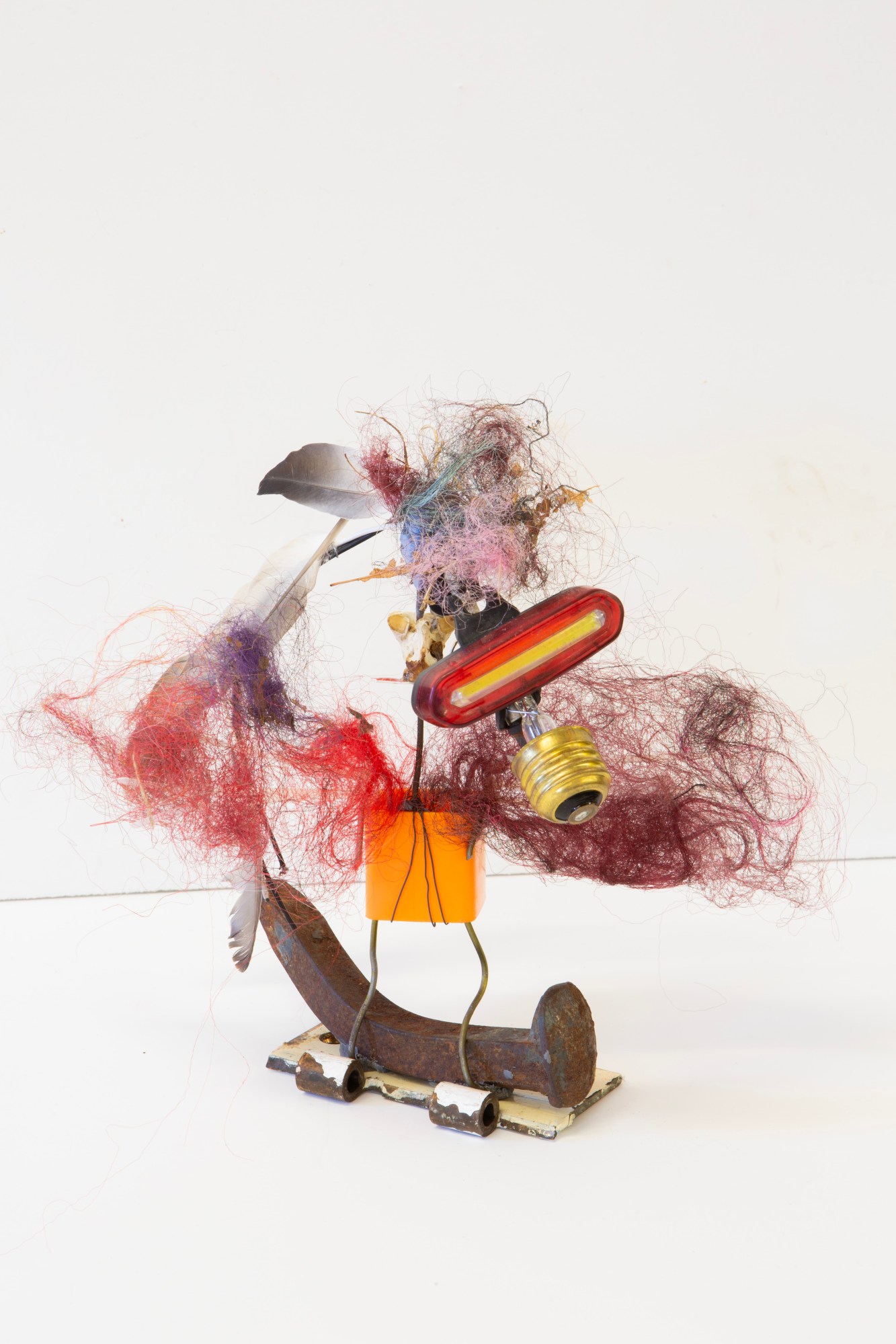 an abstract bird sculpture made from a large bent nail, wool, feathers, a light bulb and other found objects