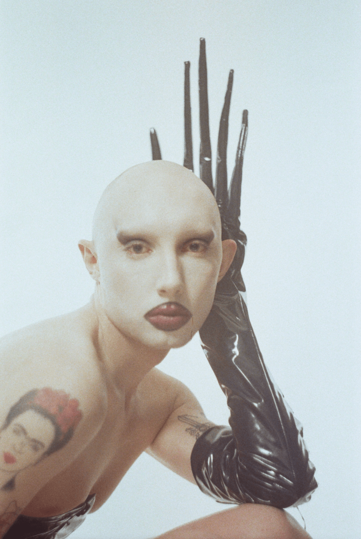 a headshot of gena marvin wearing bold makeup and latex opera gloves with long talons; they have a frida kahlo tattoo on their arm