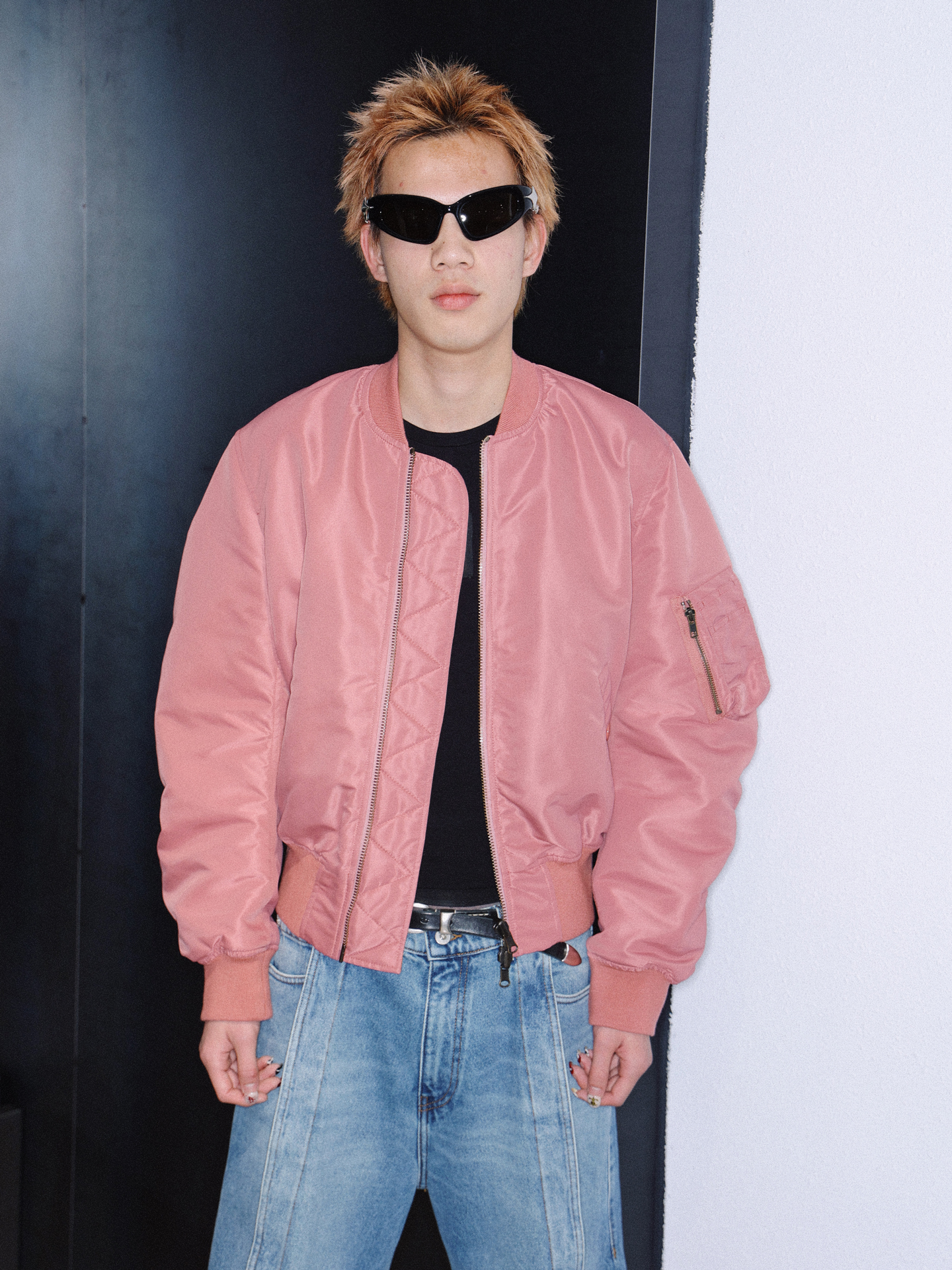 a man with spiky bleached hair wears 00s sunglasses with a pink bomber jacket and stonewash jeans