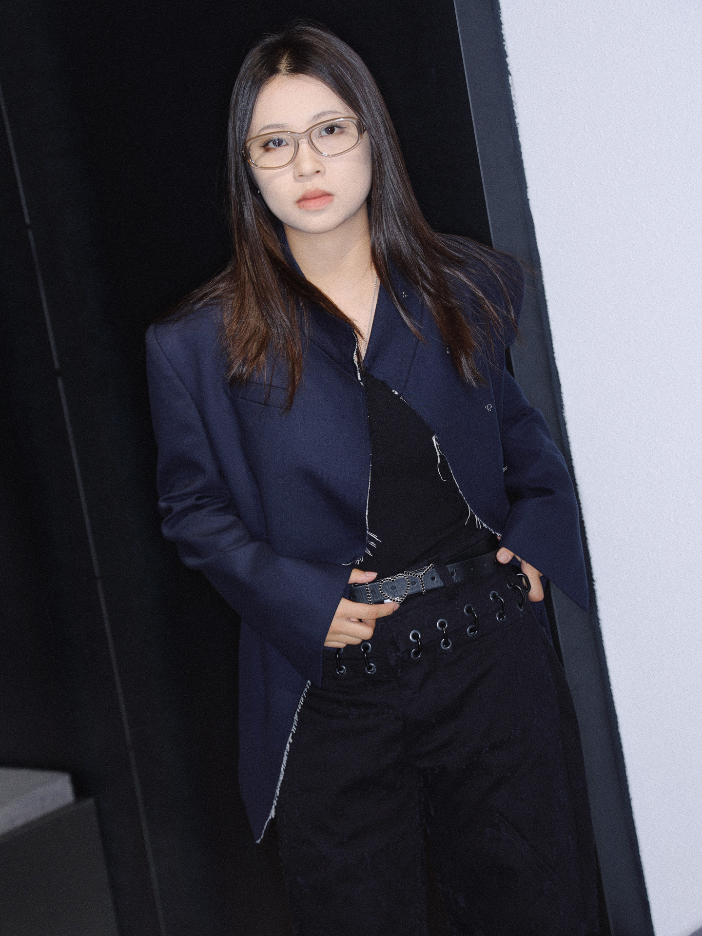 a woman in glasses and an oversized navy blazer holds her belt and looks to camera