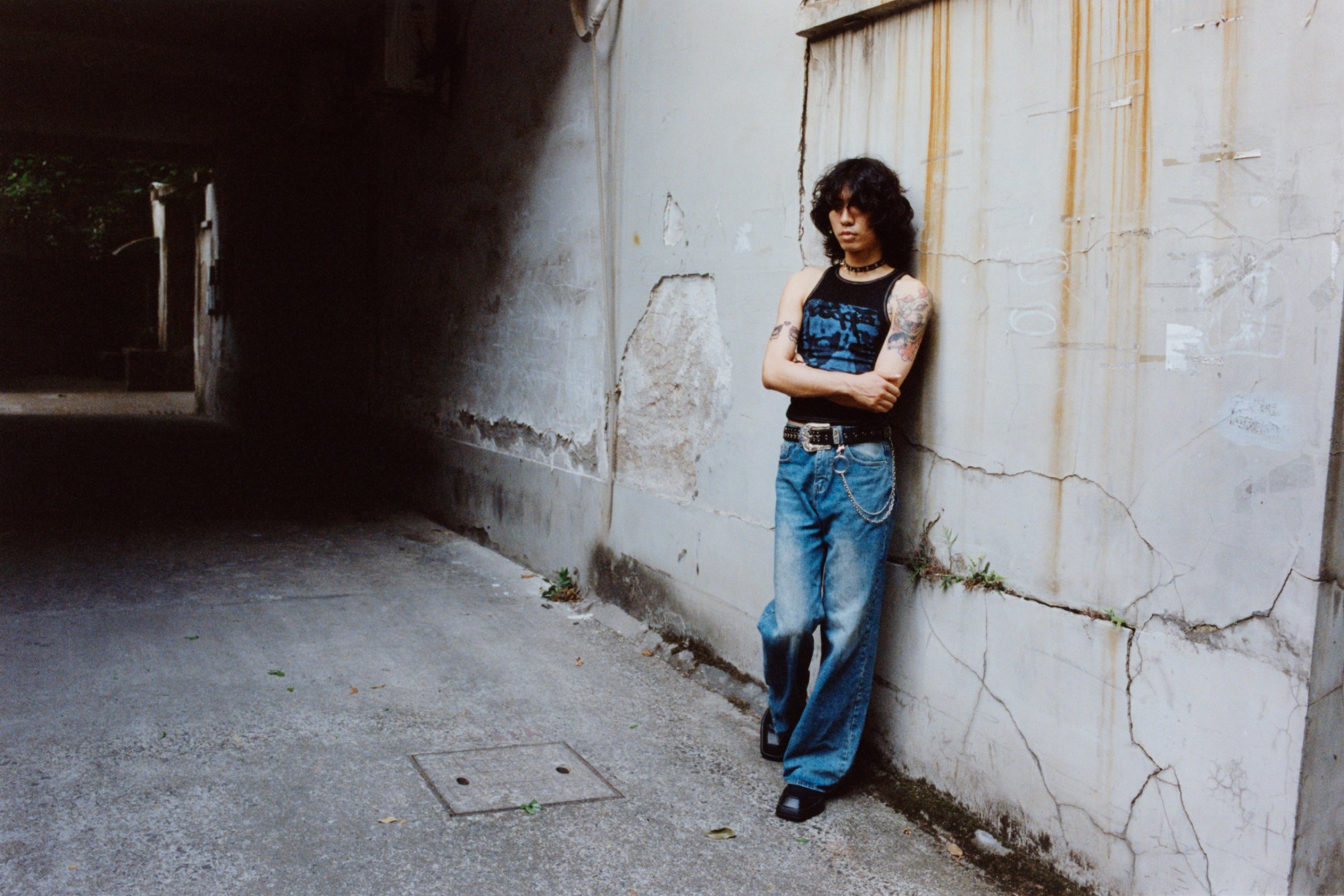 a young man in a black sleeveless top and blue jeans stands against a grey concrete wall