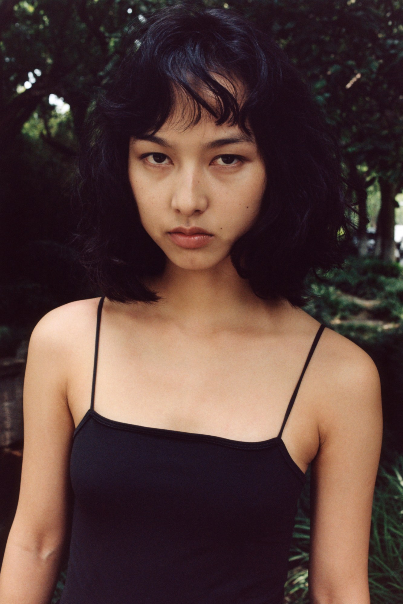 a young woman with short black hair in a black tank top standing in front of greenery