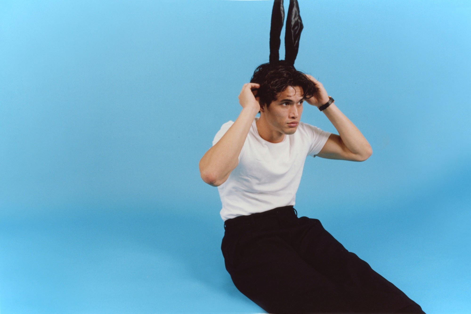 charles melton sitting against a blue studio backdrop wearing a pair of black bunny ears