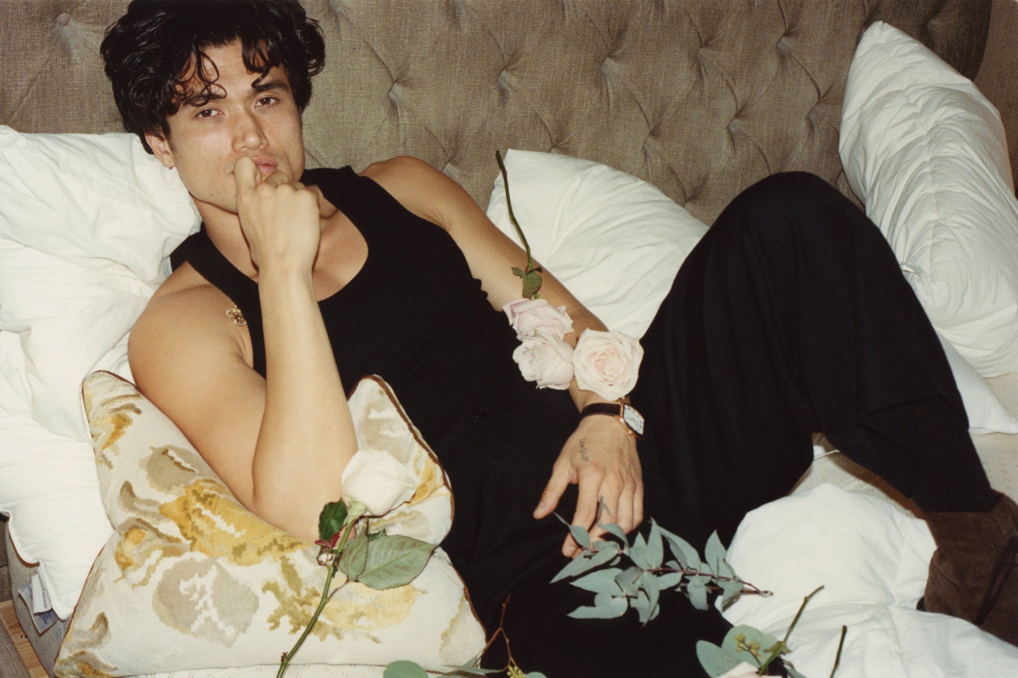 charles melton in a black tank top, reclining on a hotel bed surrounded by roses