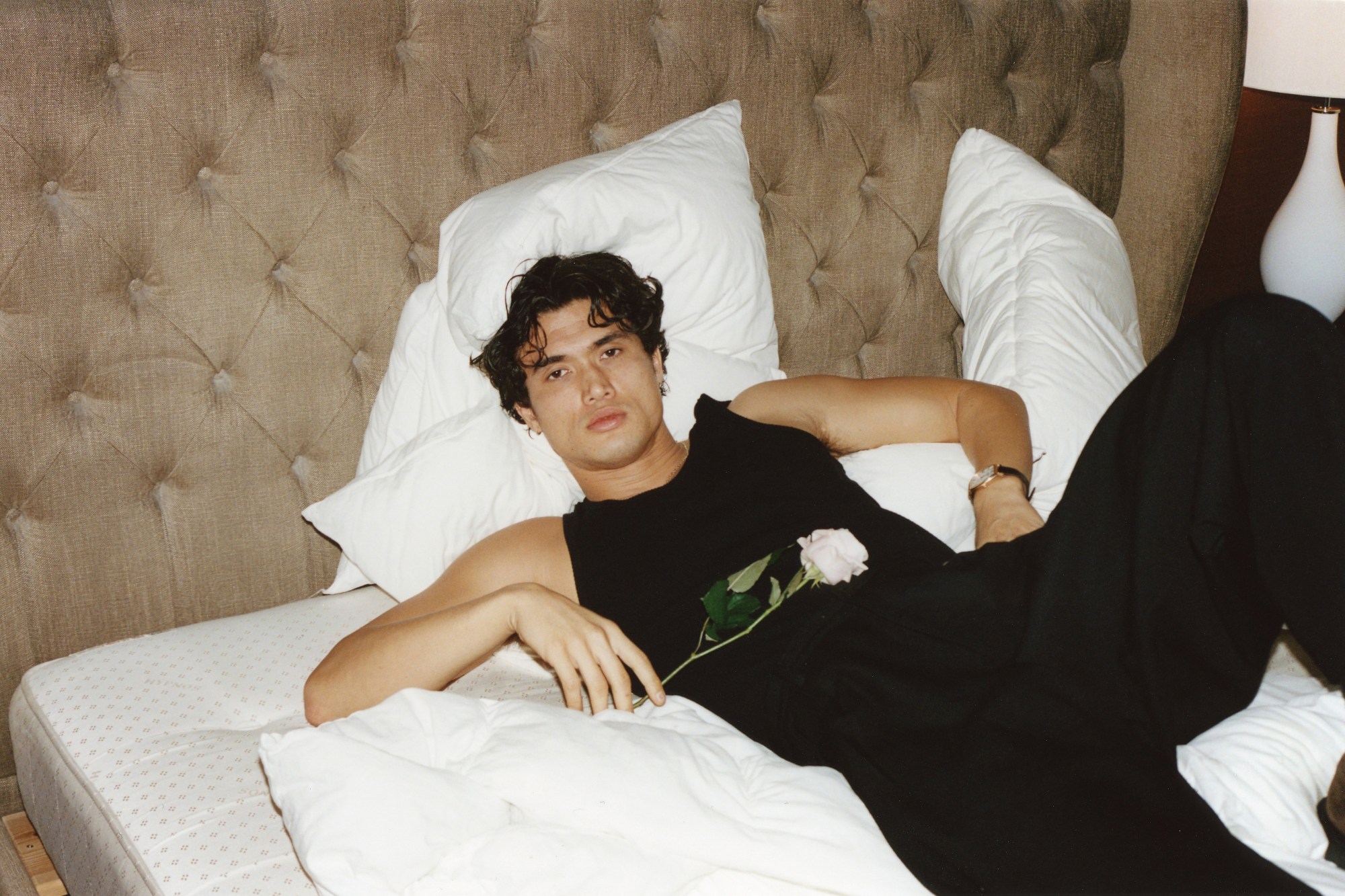 charles melton reclining on a hotel bed holding a single white rose