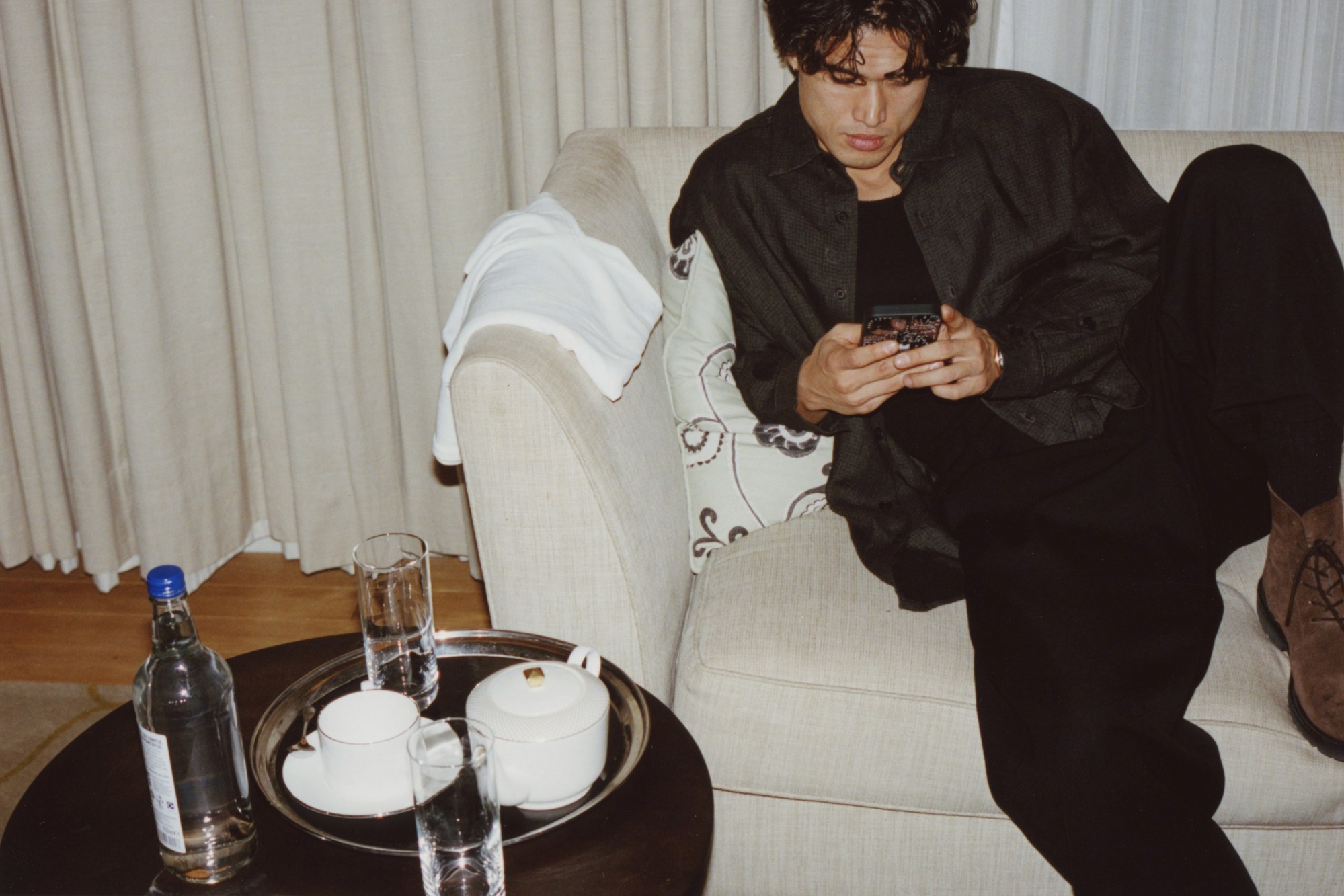 charles melton wearing all black, sitting on a hotel room sofa scrolling on his phone