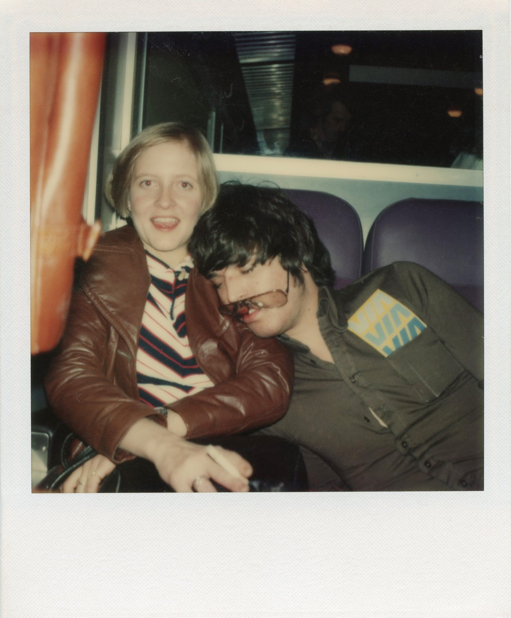 a blonde woman in a brown leather jacket sits at the back of a tour bus, a man in a tight shirt and aviators is falling asleep on her shoulder