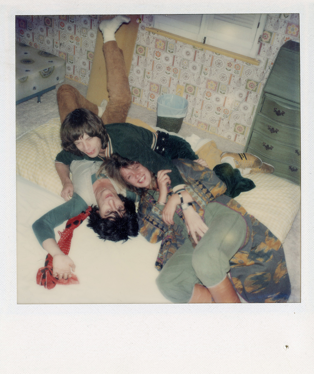 mick jagger, ronnie wood and a friend playfully pile on top of each other on a yellow plaid mattress on the floor of a bedroom