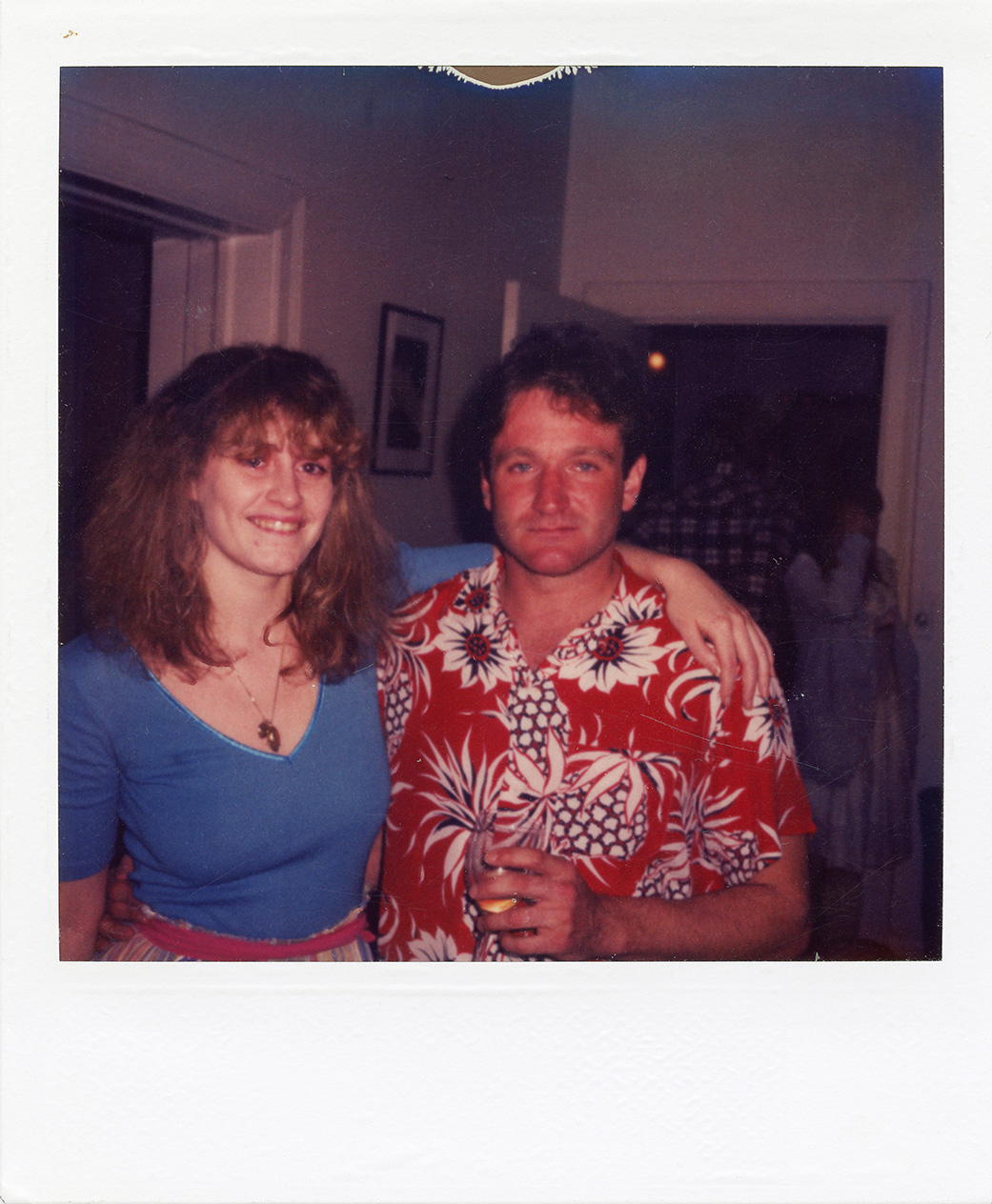 robin williams (wearing a red hawaiian shirt) and a female friend smile for a photo at a house party