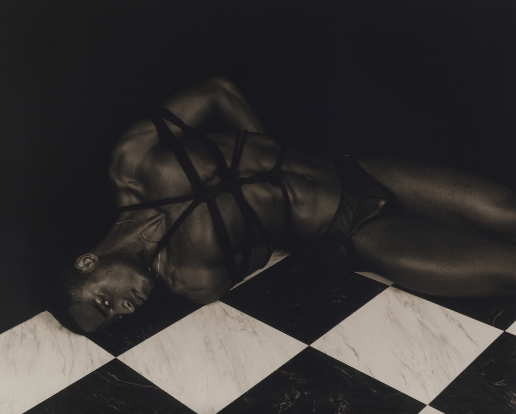 Photograph by Quil Lemons of a man in briefs and tied up lying in the corner of a room with a checkered floor from Quiladelphia