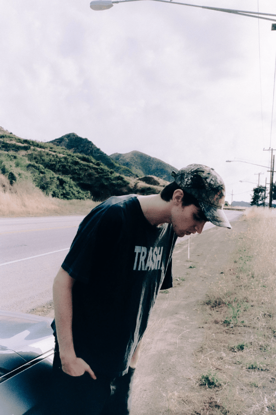 Photograph by Saem of a friend he made online, standing by their car wearing a top saying trash and spitting on the ground